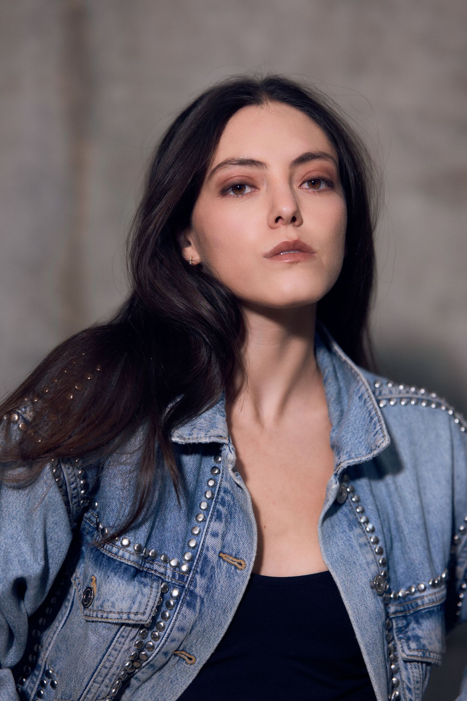 Young woman with dark hair wearing a studded denim jacket over a black top, posing confidently indoors.