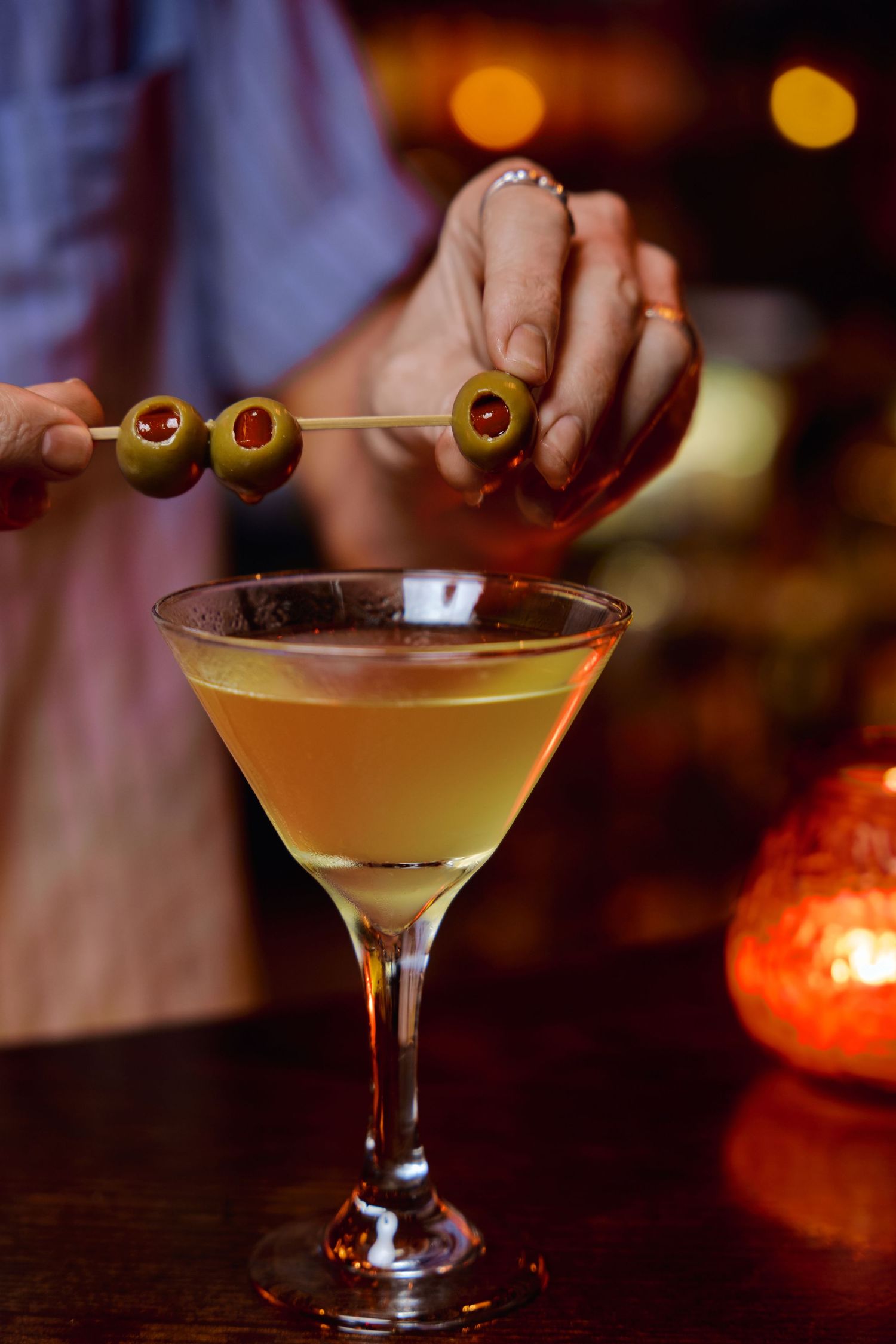 Hands garnish martini cocktails with olives in dimly lit bar setting.