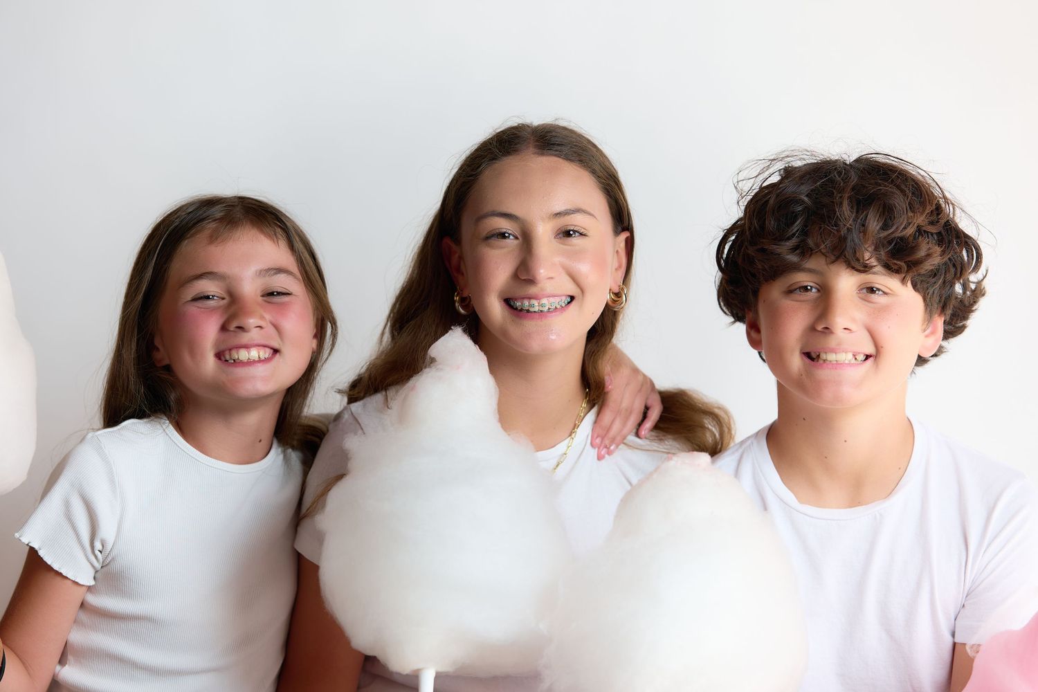 Three smiling kids holding white cotton candy against a white background, wearing matching white t-shirts.
