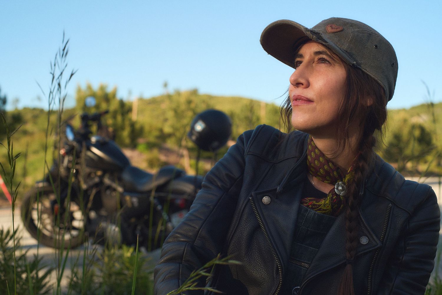Woman in cap and leather jacket sits outdoors near a motorcycle, gazing into the distance on a sunny day.