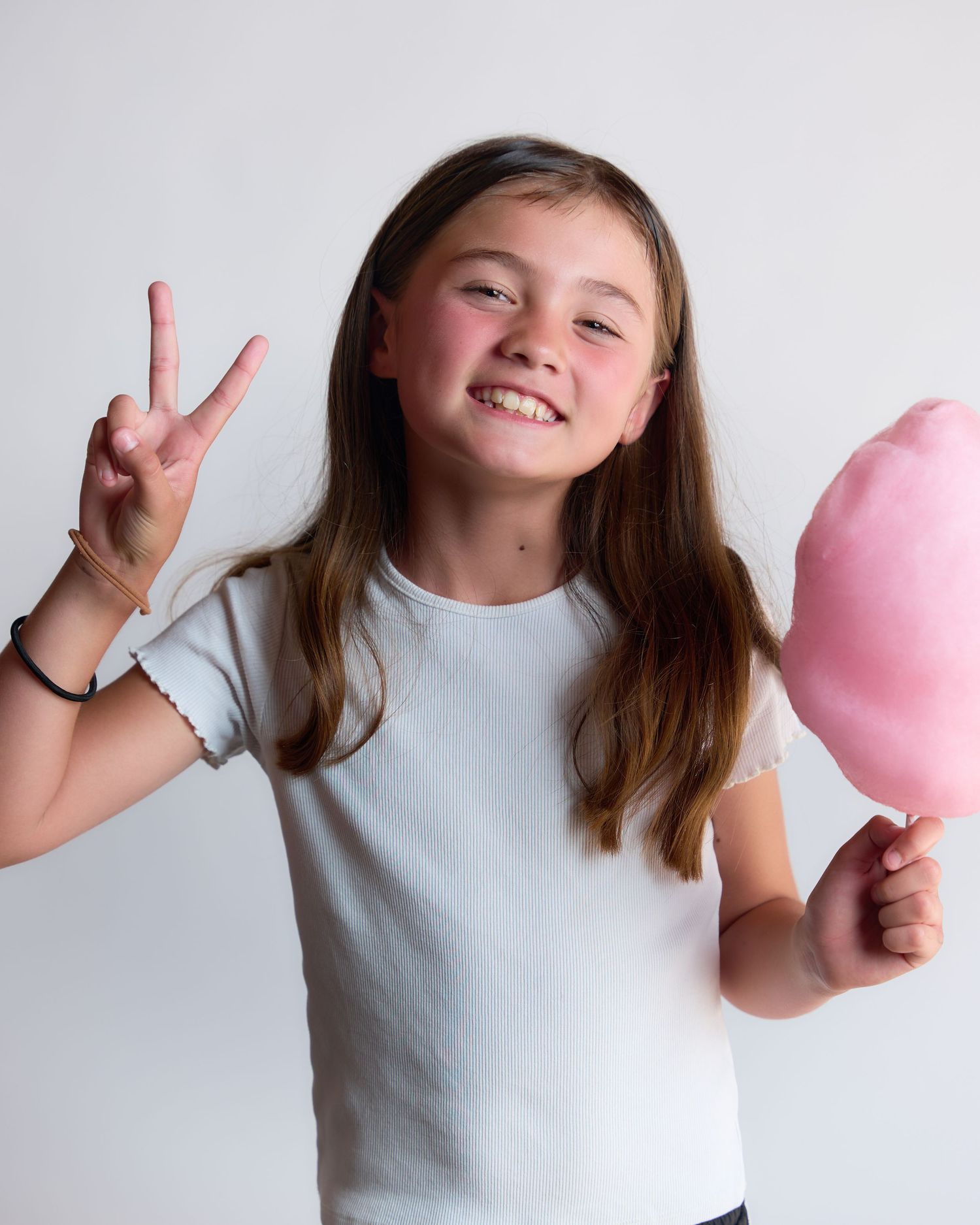 Smiling girl holding pink cotton candy and making a peace sign, wearing a white t-shirt.