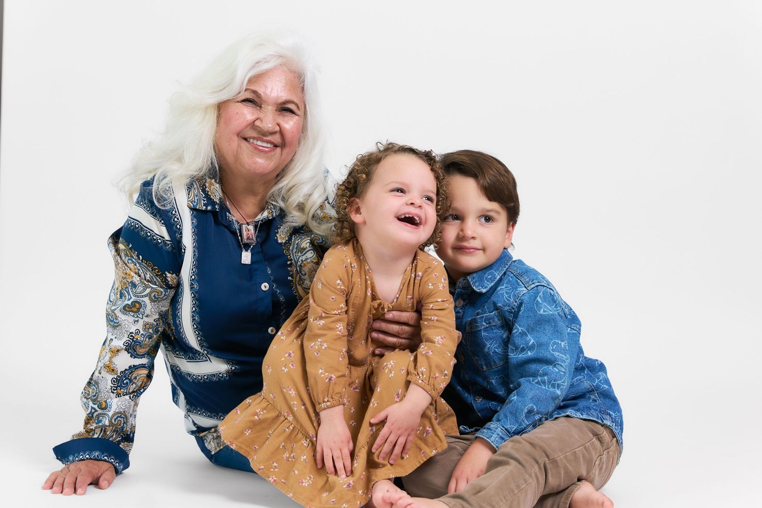 Elderly woman with white hair smiles warmly with two young children in a bright studio portrait.