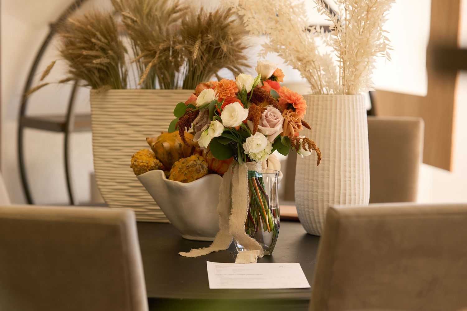A vase of orange and white roses sits on a dining table next to a bowl of fruit and white ceramic decor.