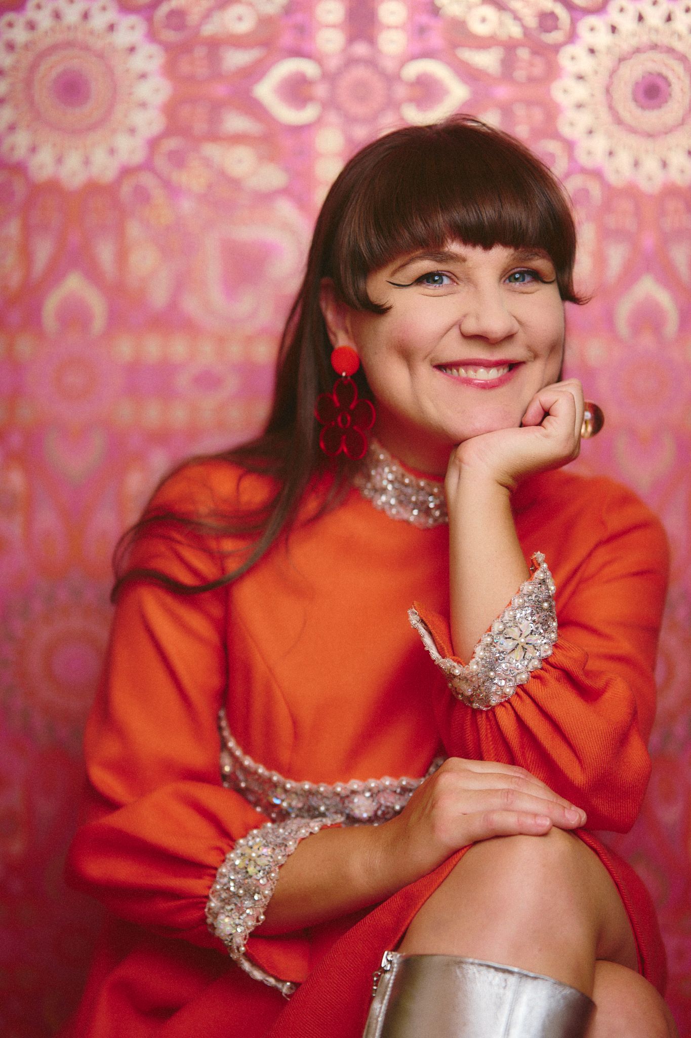 Person in bright red outfit with sparkly accessories poses against pink patterned background.