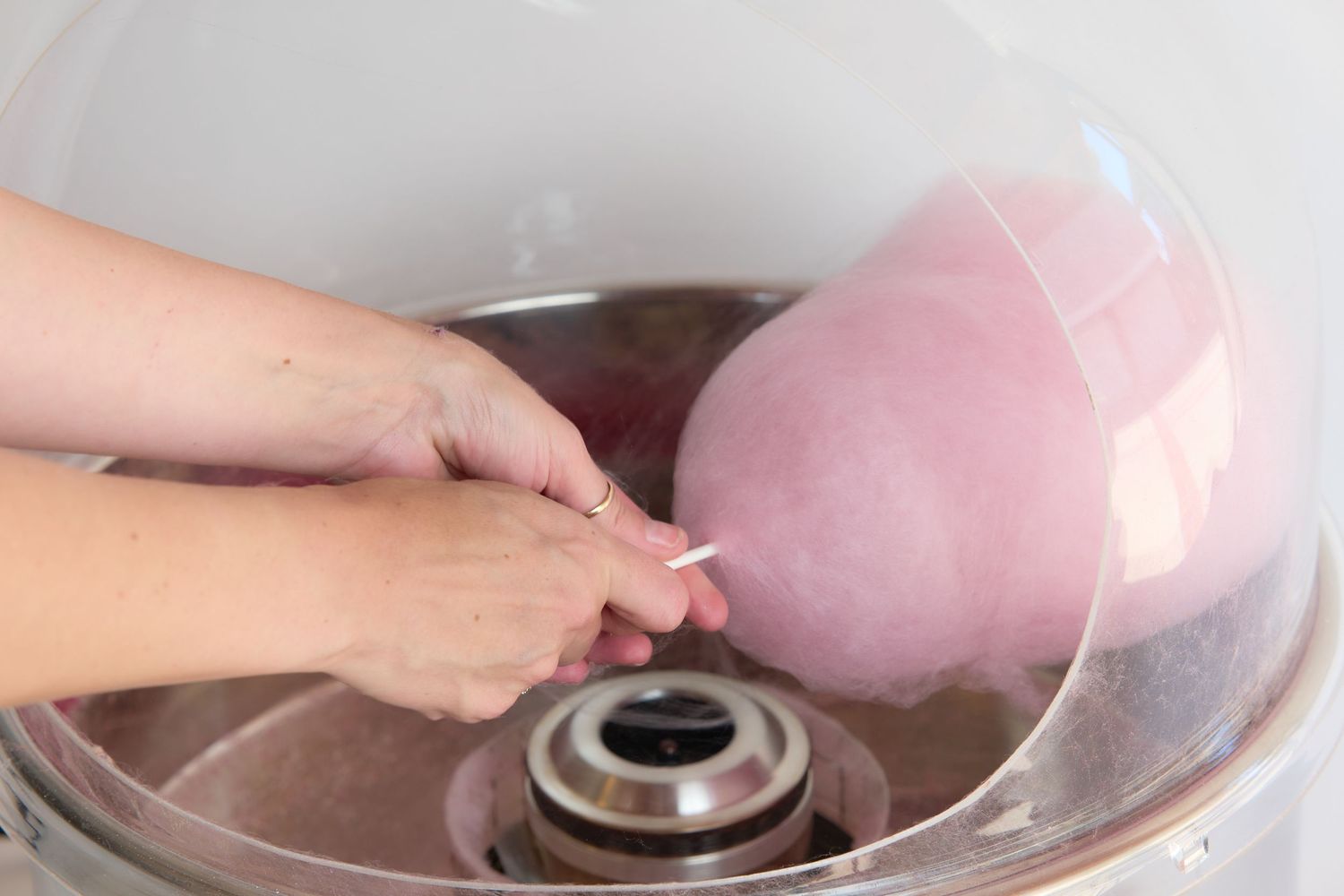 Hands spinning pink cotton candy on a machine with a clear bowl in a close-up view.