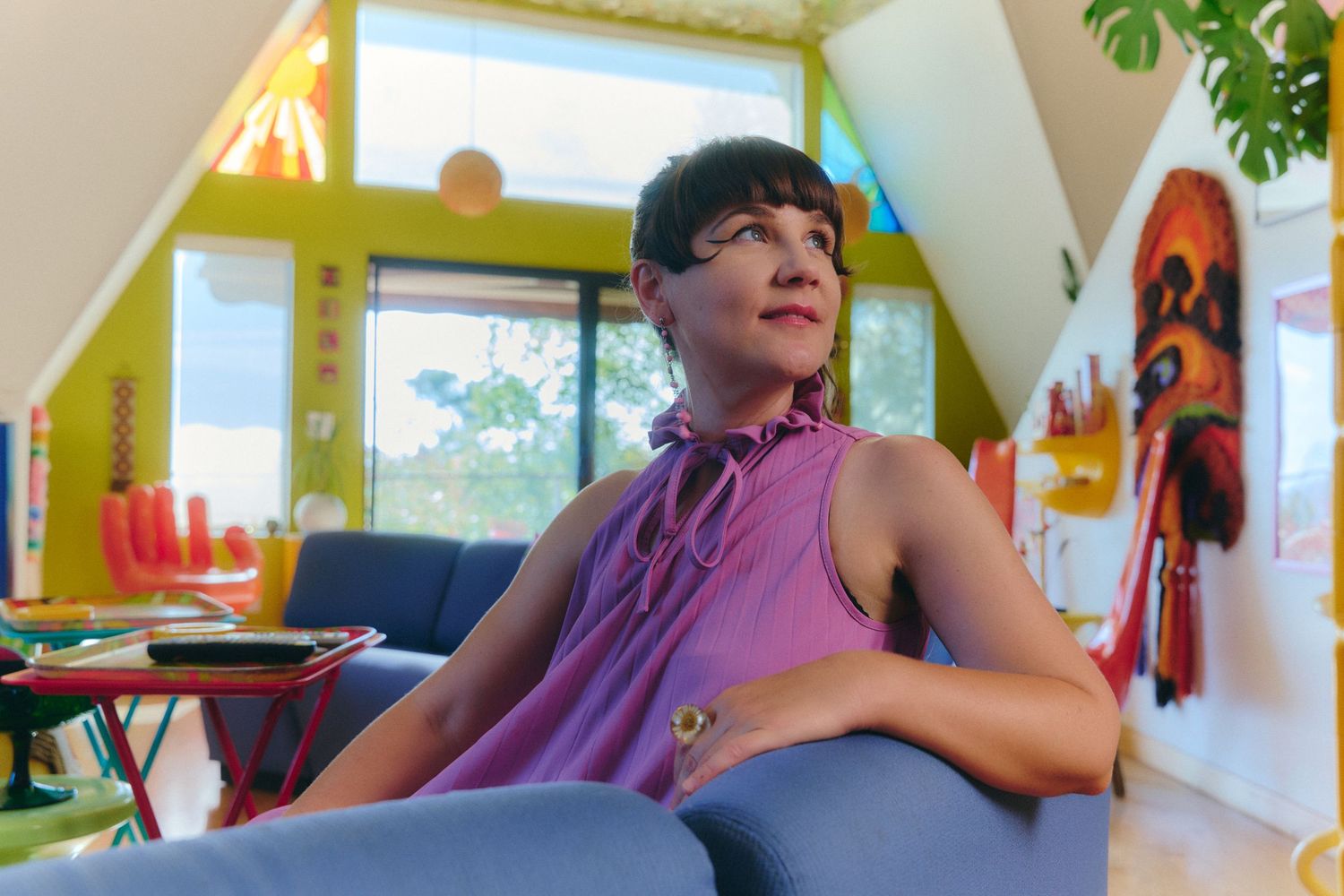 Woman in purple sleeveless top relaxing in colorful artsy room with bright yellow walls and eclectic decor.