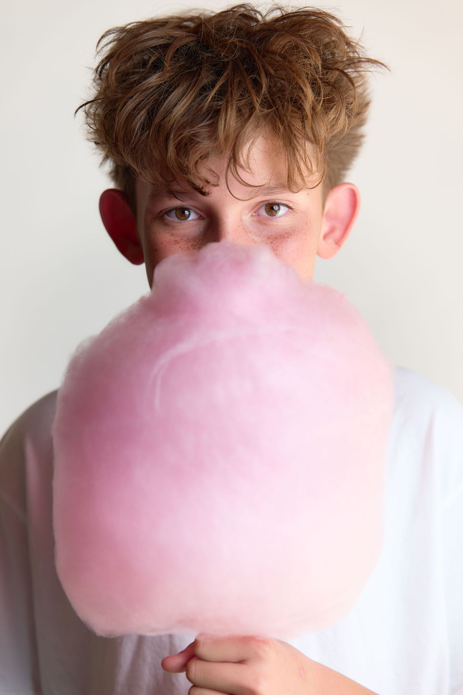 A young boy holds pink cotton candy covering the lower half of his face against a white background.