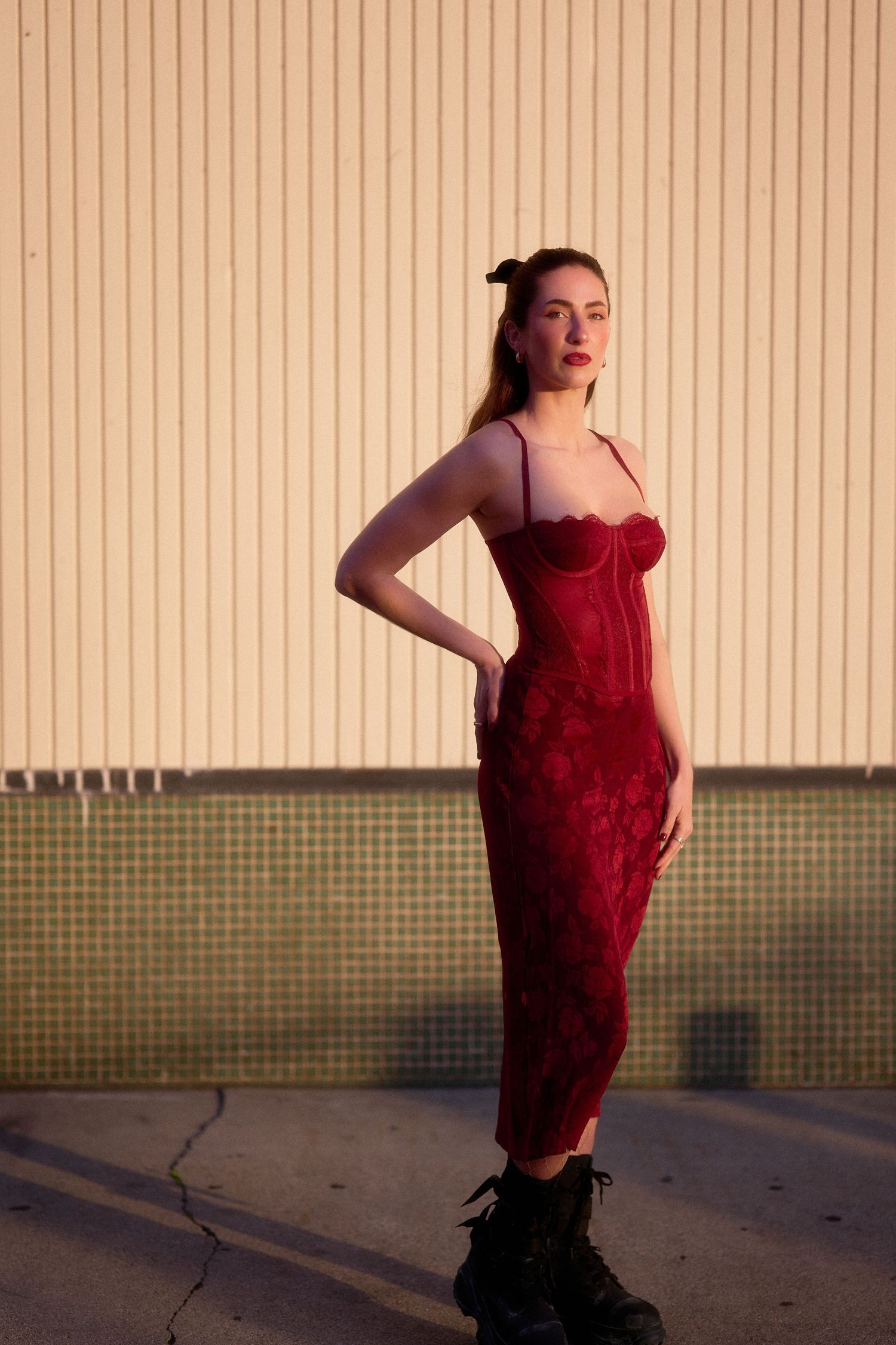 Model in red lace dress posing against beige striped wall at sunset.