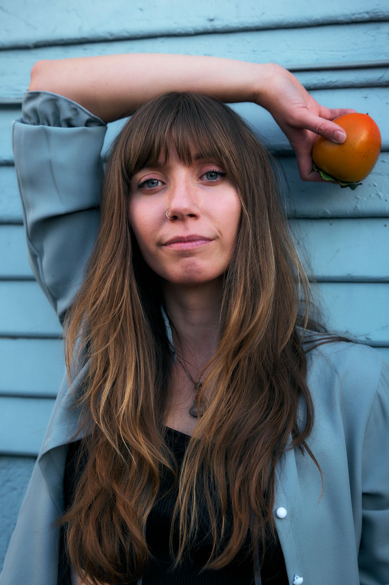 Young woman with long brown hair holding a tomato against a blue wall, wearing a gray jacket and necklace.