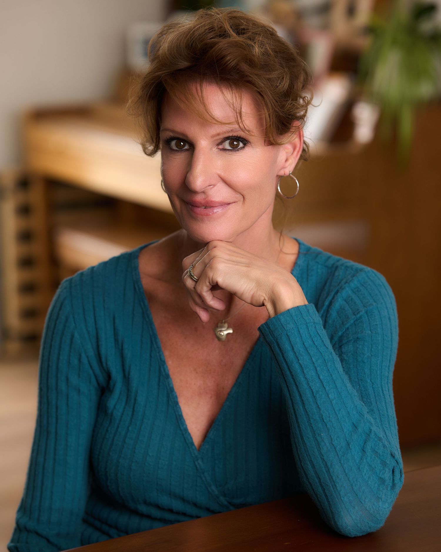 Woman in teal sweater smiling warmly, posing with hand on chin in a warmly lit indoor setting.