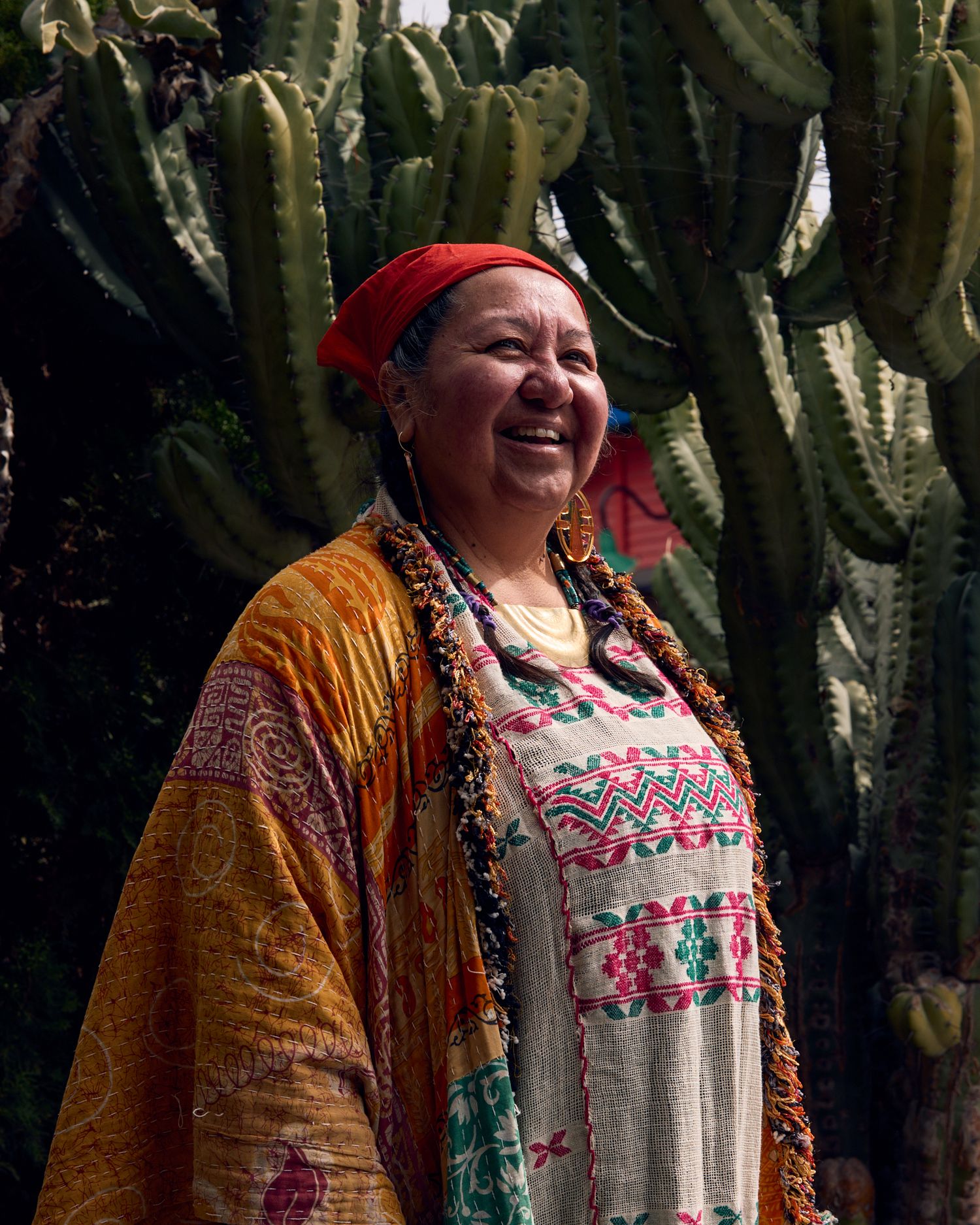 Person wearing colorful embroidered dress and orange shawl standing in front of cacti.