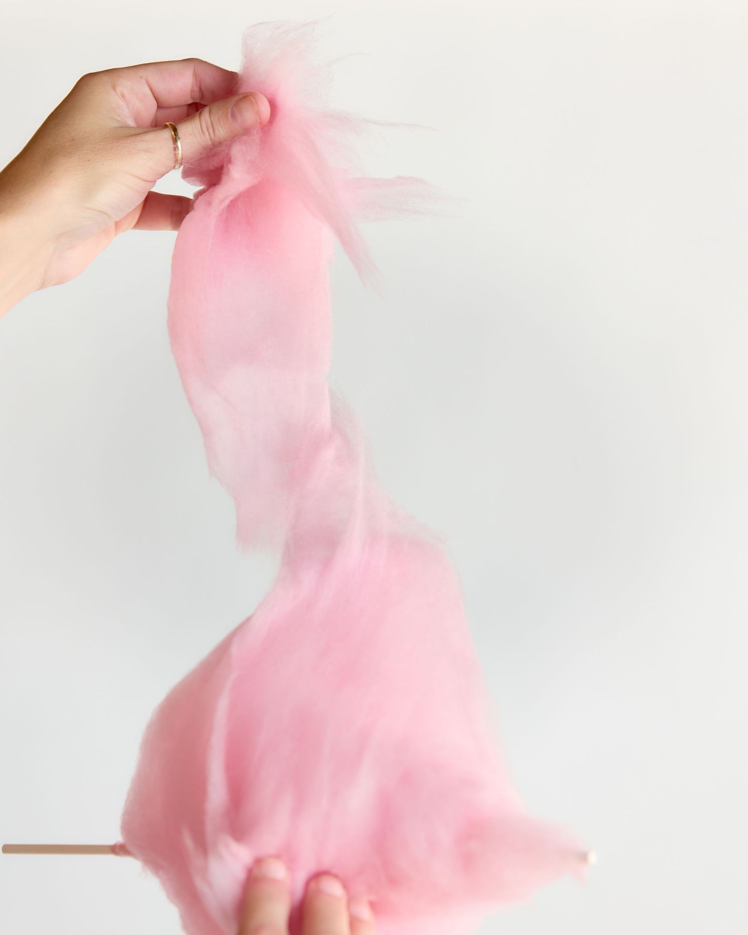 Hands stretching a large, fluffy pink cotton candy on a stick against a white background.