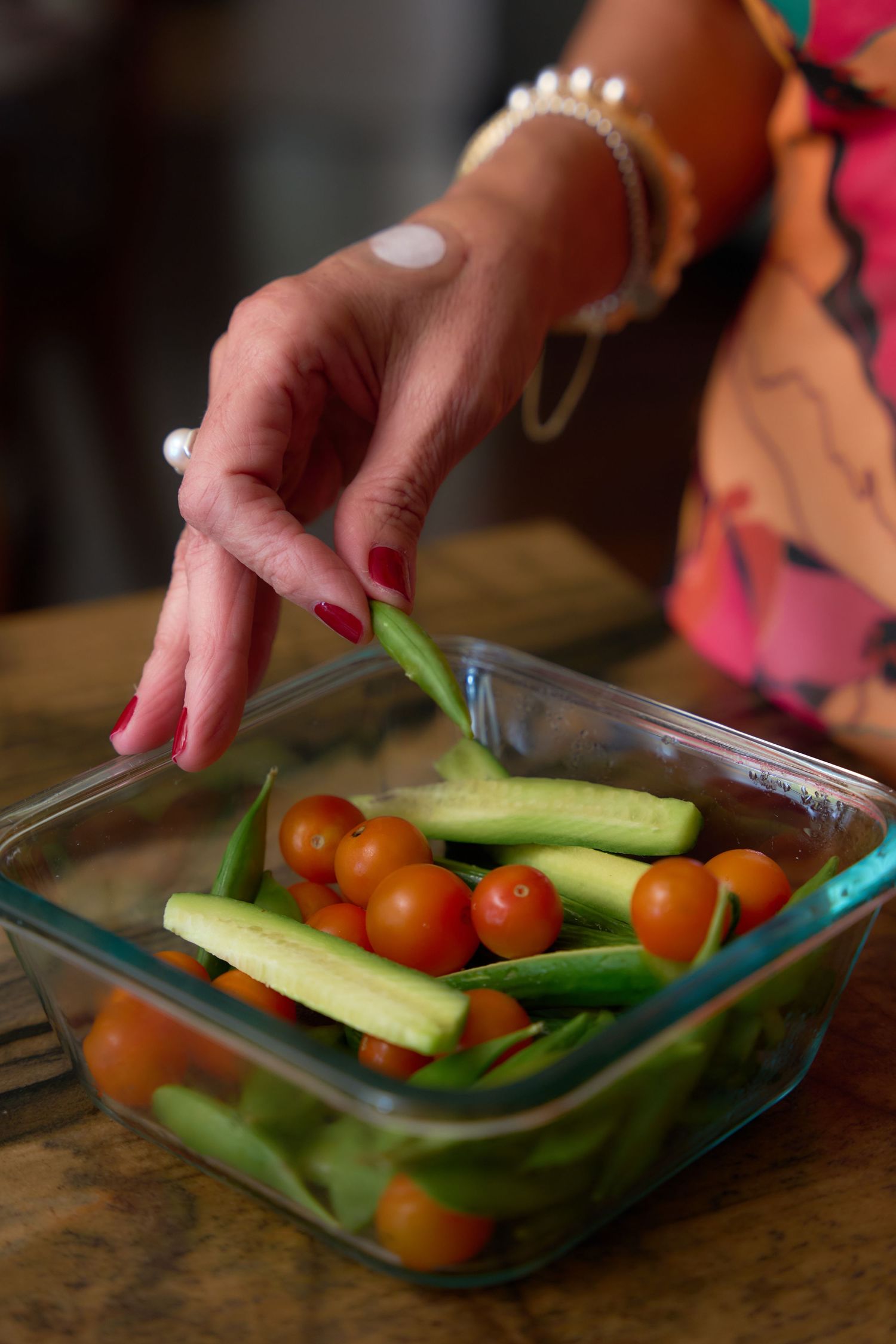 A sequence of hands placing fresh vegetables including cucumbers and cherry tomatoes into a glass meal prep container.