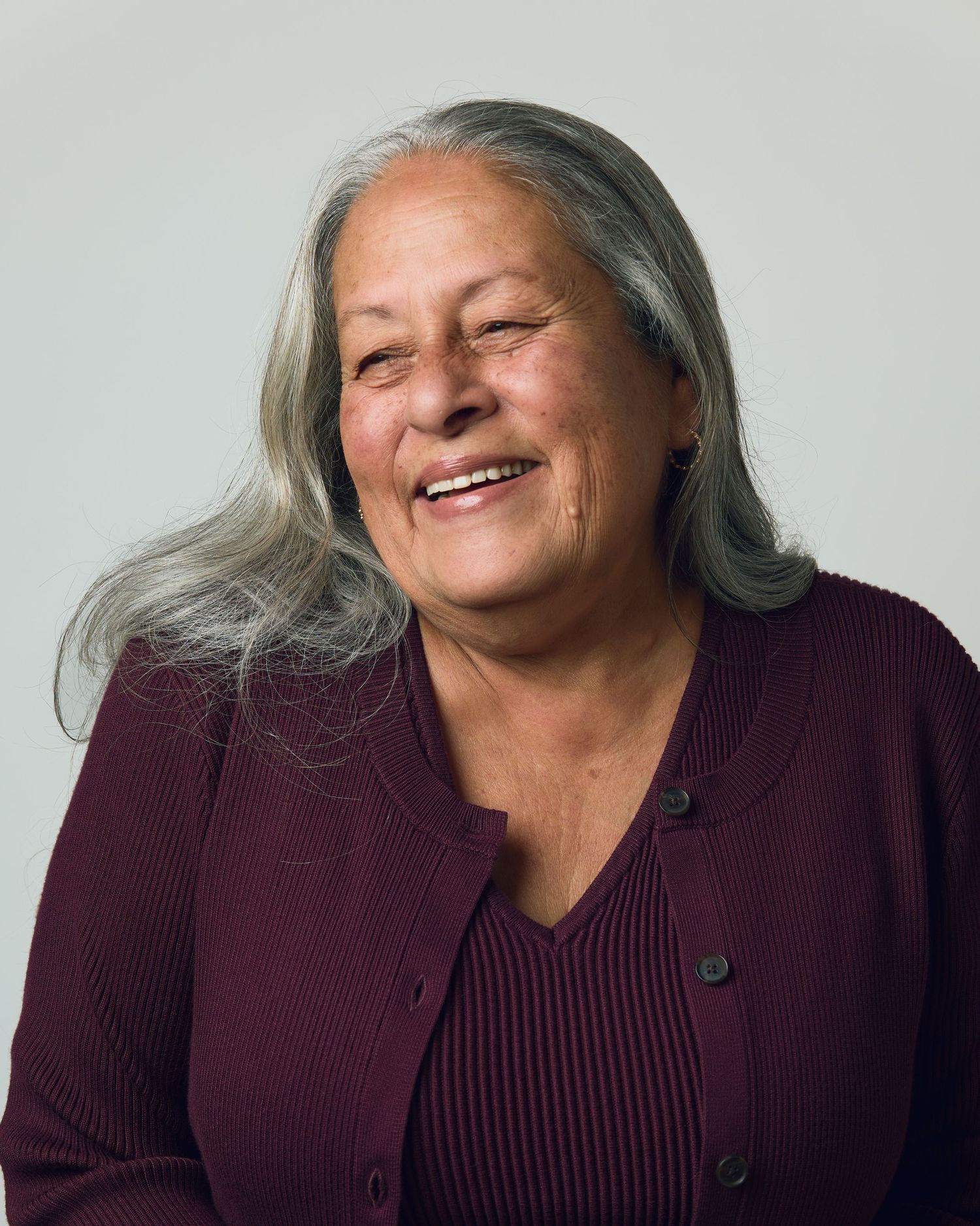 Smiling older woman with long gray hair wearing a dark red cardigan, portrait photo on light gray background.