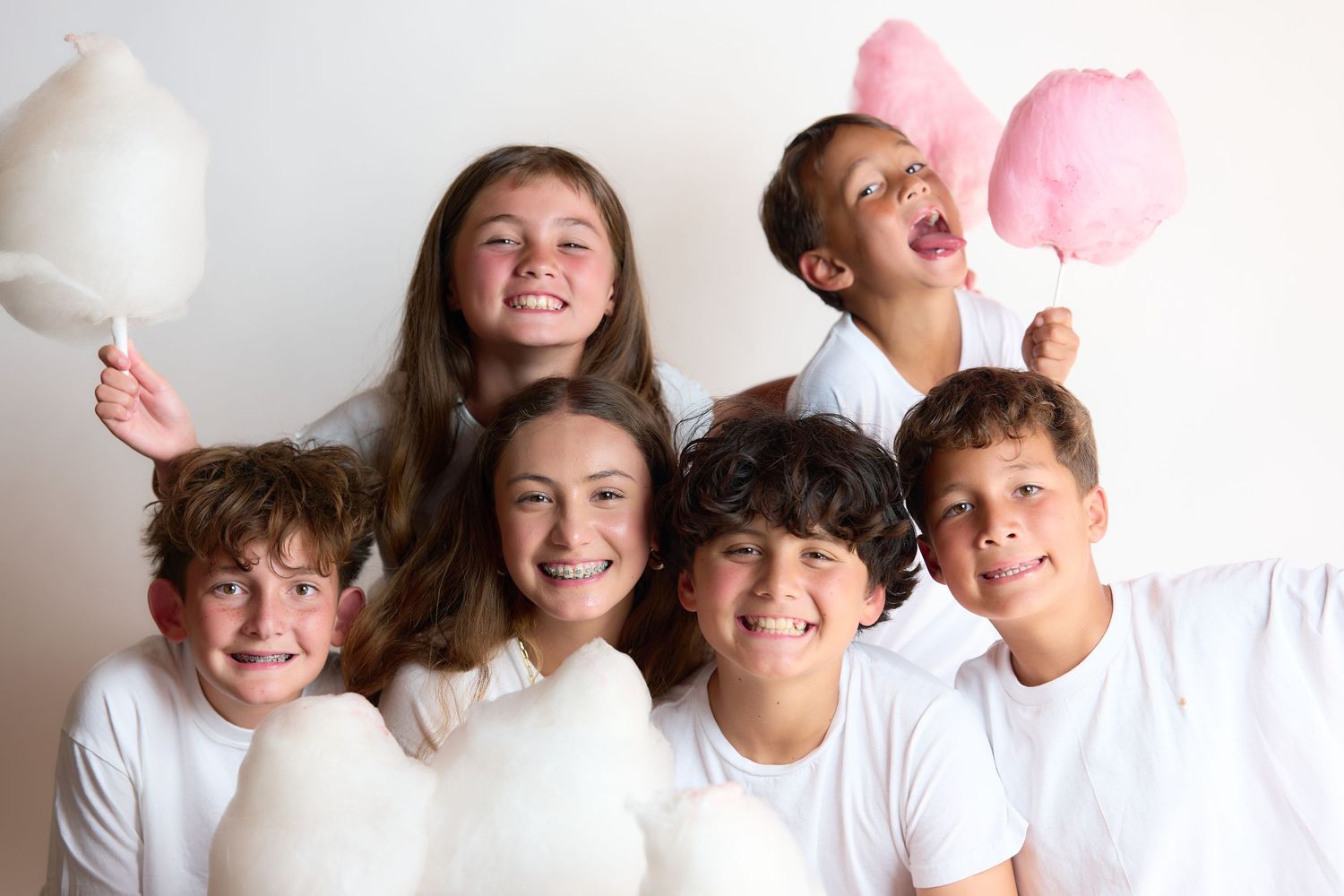 Six happy kids in white shirts holding fluffy white and pink cotton candy, smiling together against a white background.
