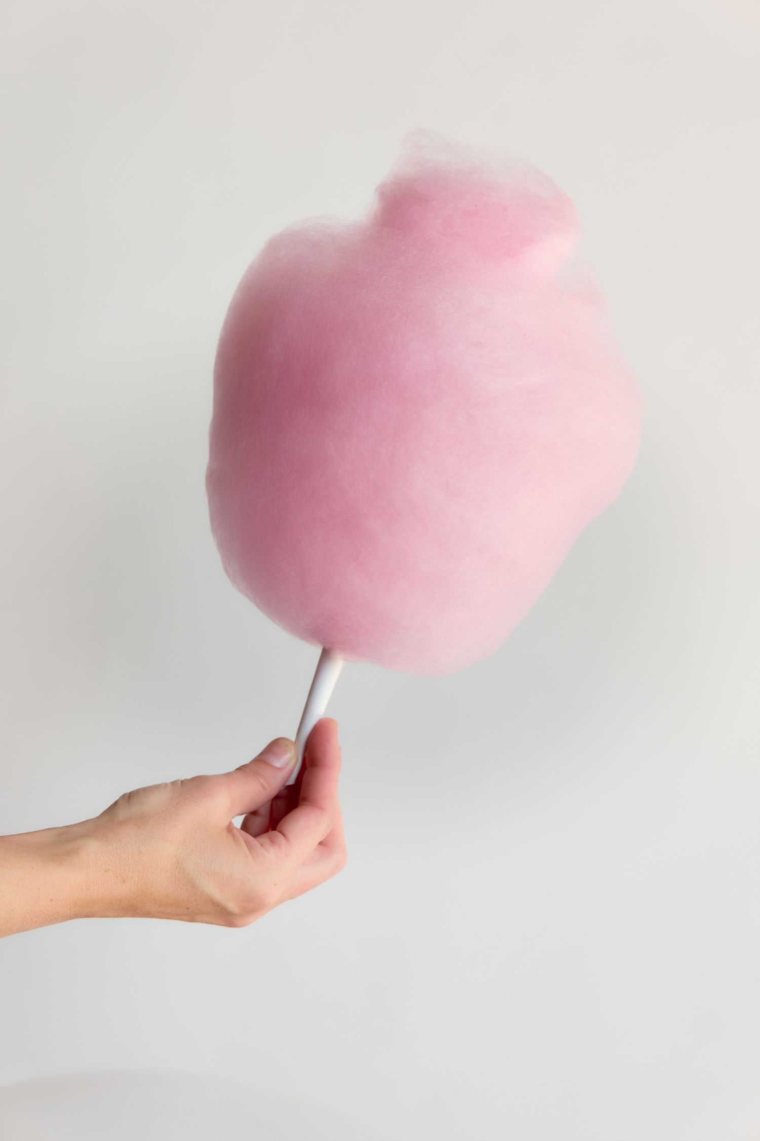 Hand holding a fluffy pink cotton candy on a white stick against a light gray background.
