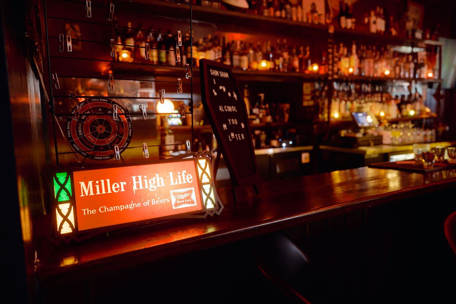Dark bar interior with illuminated bottles and Miller High Life sign glowing in warm ambient lighting.