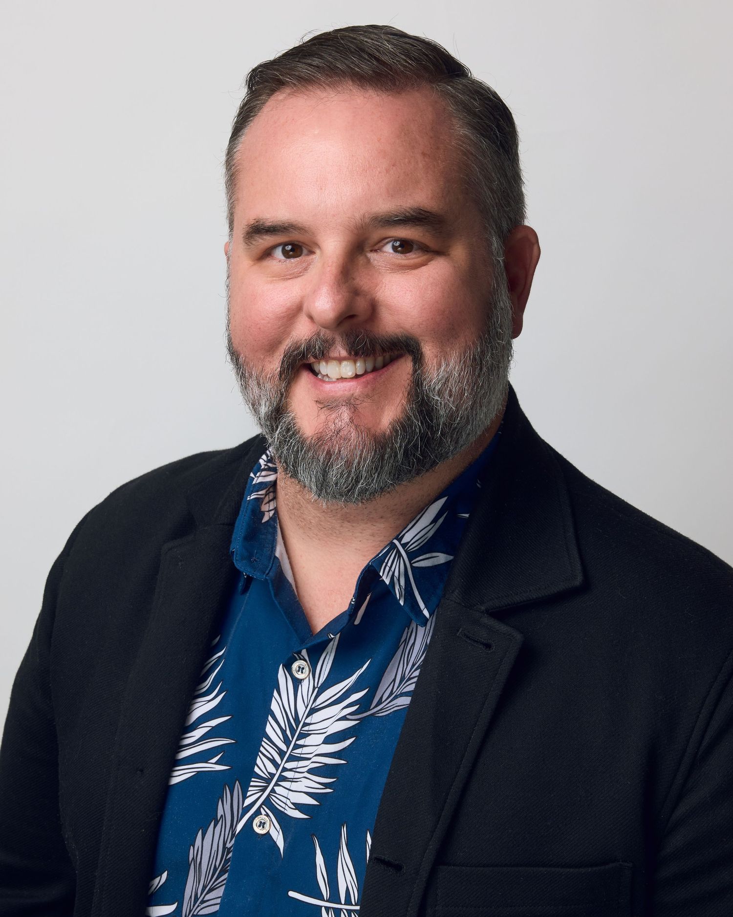 Professional headshot of someone in a black blazer and blue tropical print shirt against white background.