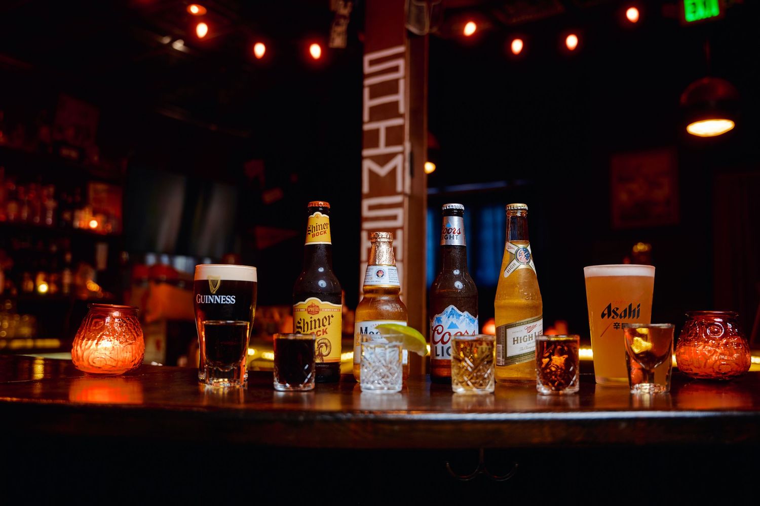 Illuminated liquor bottles glow on a dark bar shelf with red ambient lighting creating a moody atmosphere.