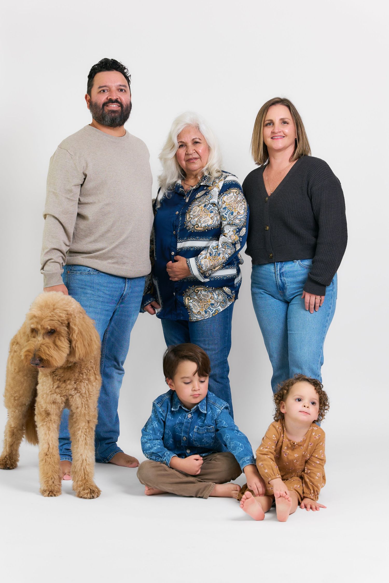 Multi-generational family portrait with two adults, elderly woman, two young children, and a golden doodle dog on white background.