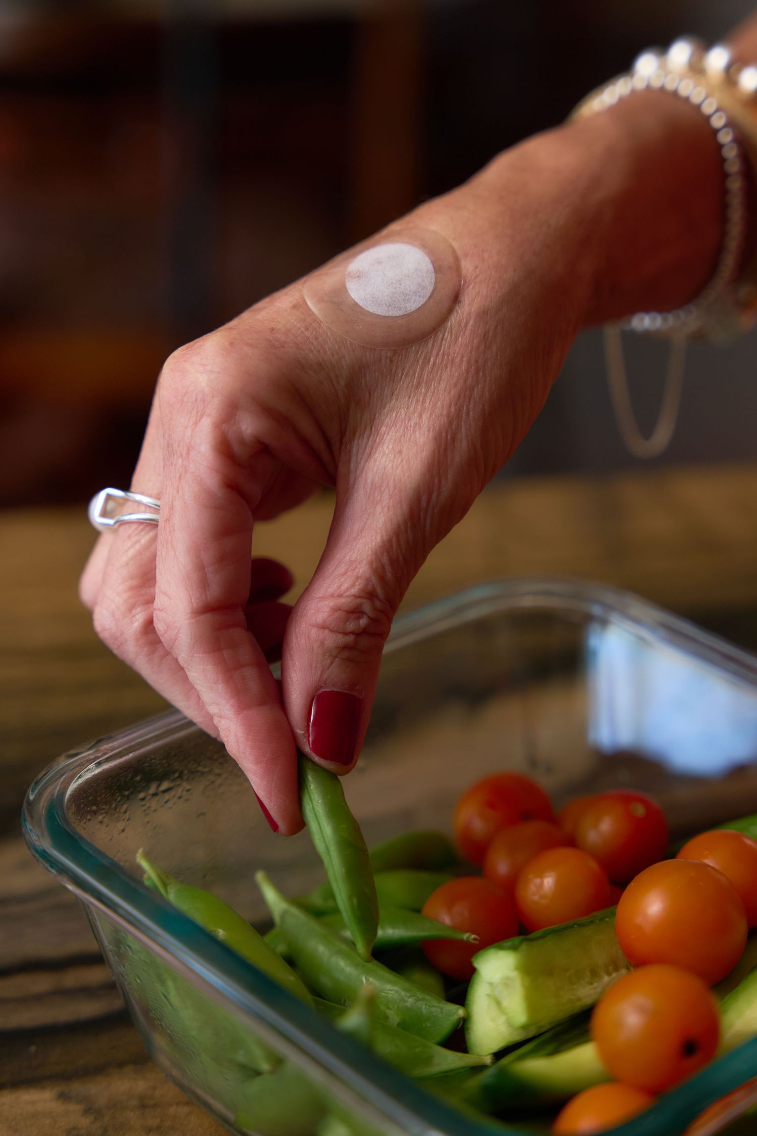 Hands placing fresh vegetables including cherry tomatoes and green bell peppers into a glass storage container.