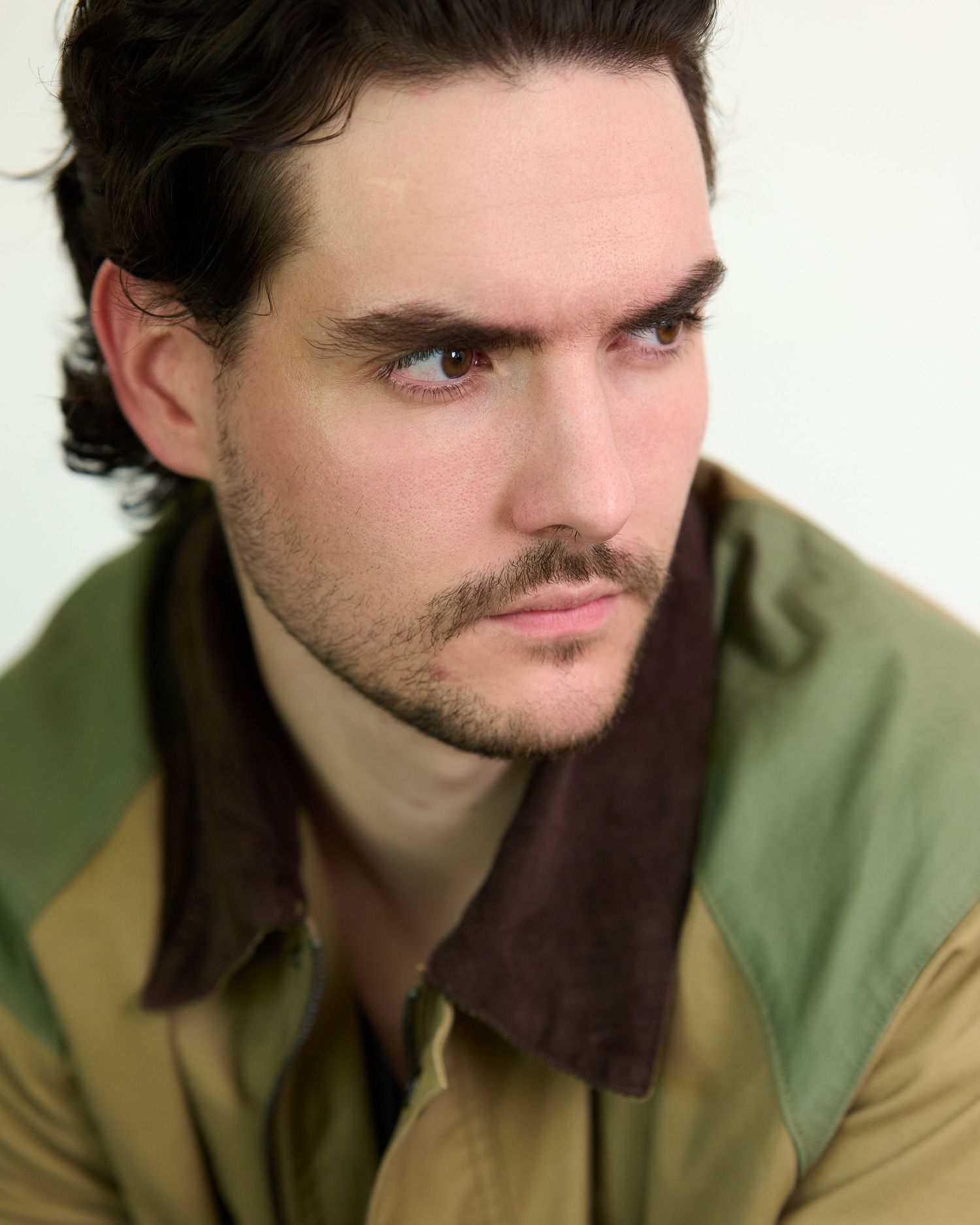 Young man with dark hair and beard wearing a olive green jacket, looking thoughtfully to the side.