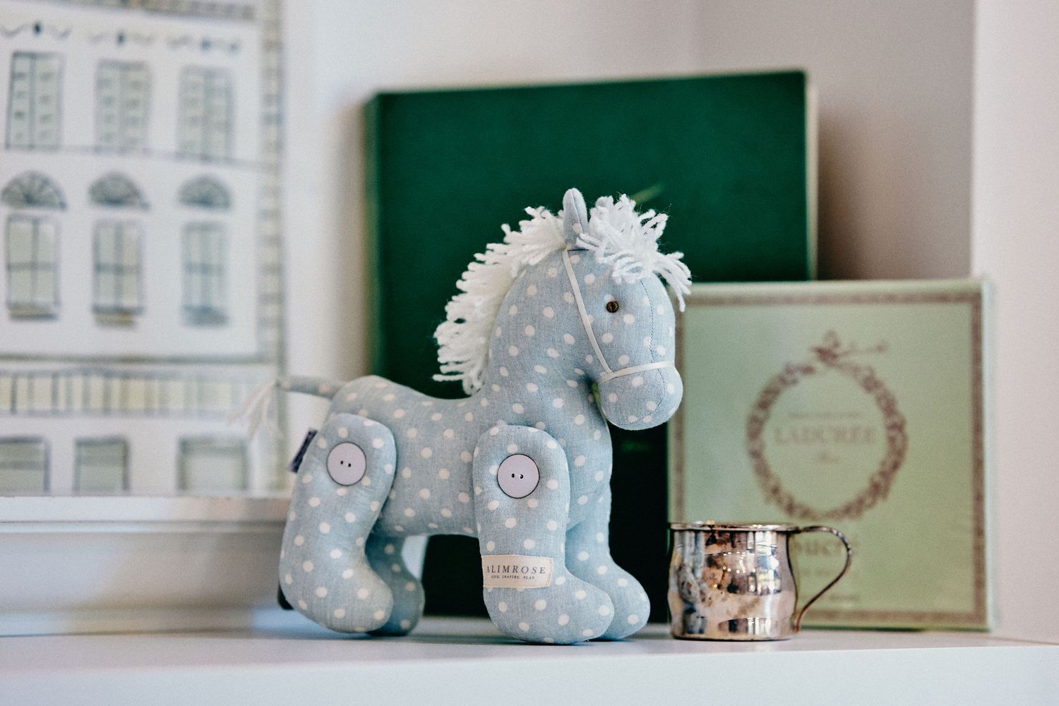 A soft blue plush rocking horse toy sits on a shelf with vintage books and a small decorative teacup.