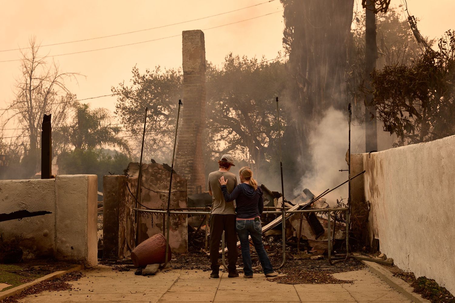 Firefighters assess damage at a burned structure with smoke rising in the background during sunset.