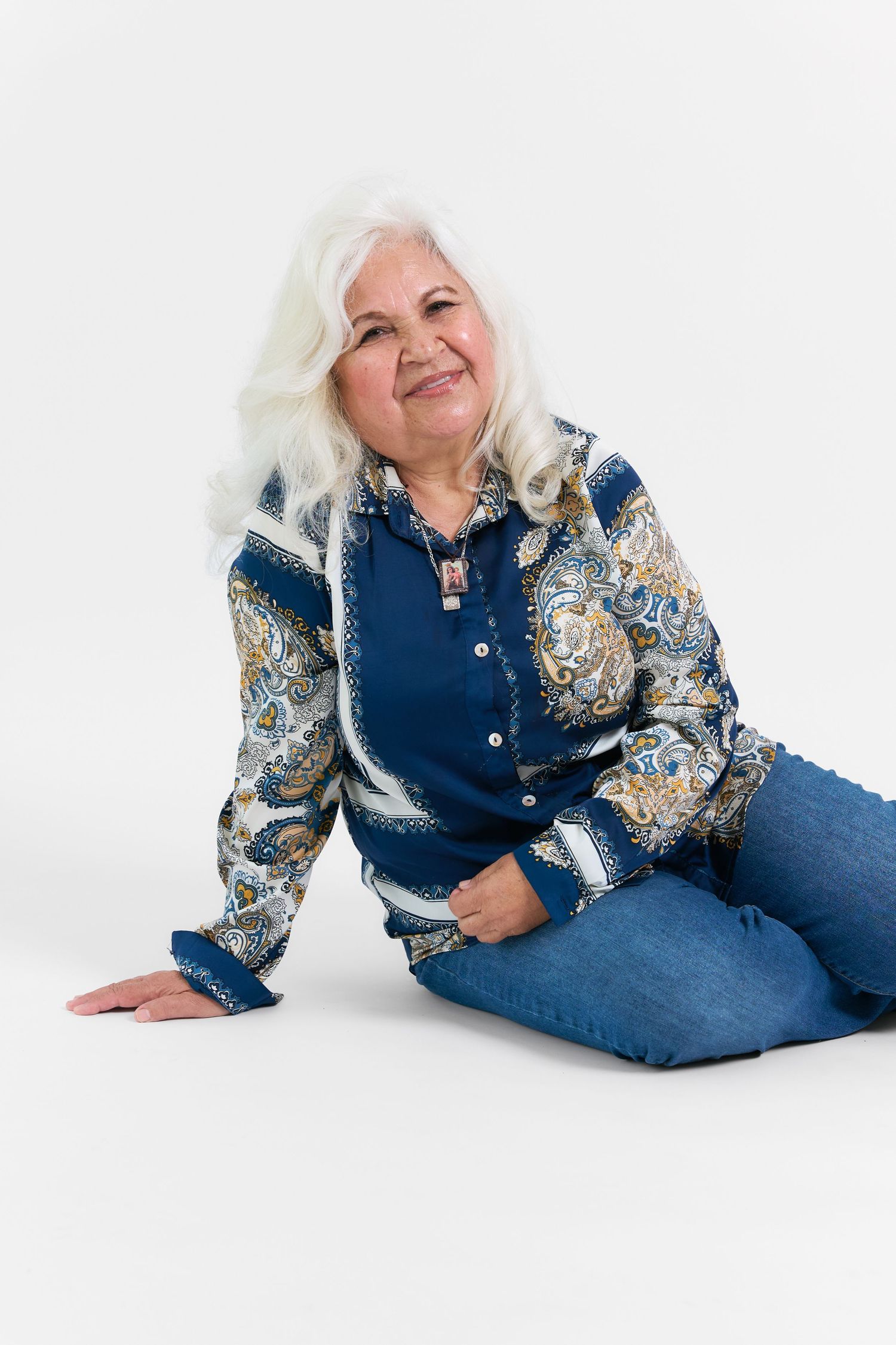 Older woman with white hair sitting on floor, wearing blue paisley blouse and jeans, smiling warmly.