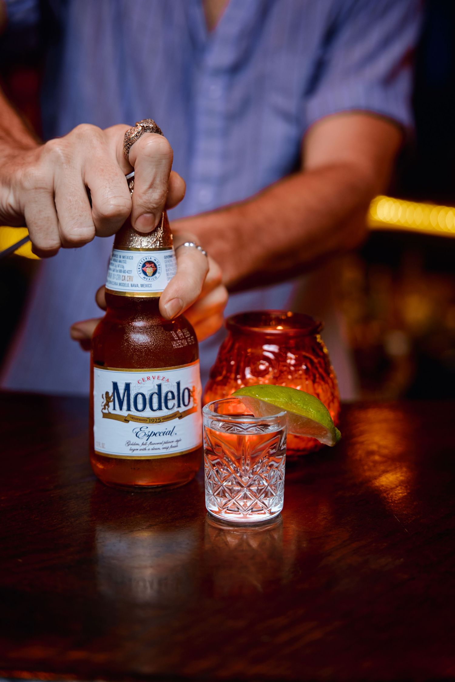 A bottle of Modelo beer being opened next to a cocktail glass and burger on a dark wooden bar counter.