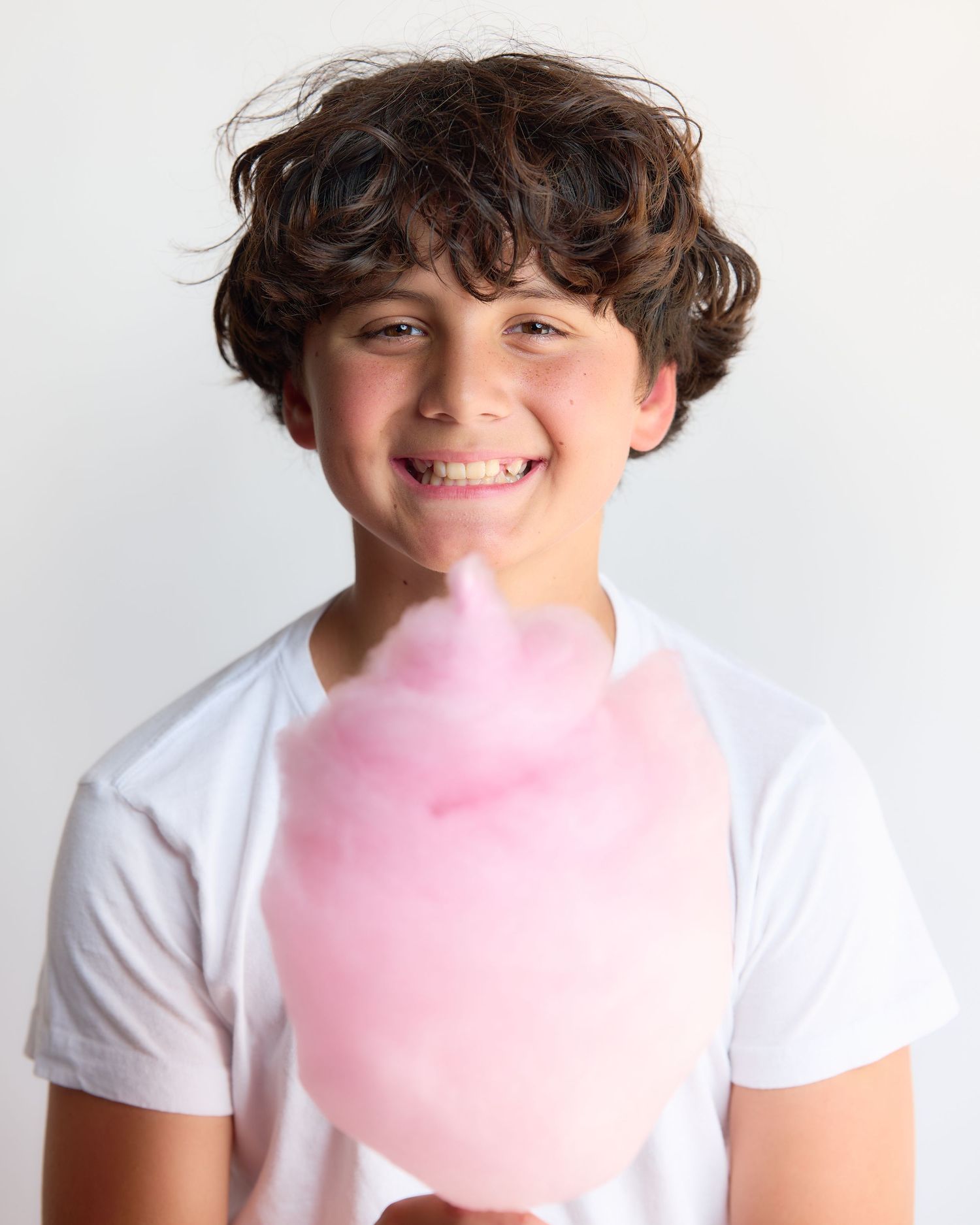 Smiling boy with curly hair holding pink cotton candy, wearing a white t-shirt against a light background.