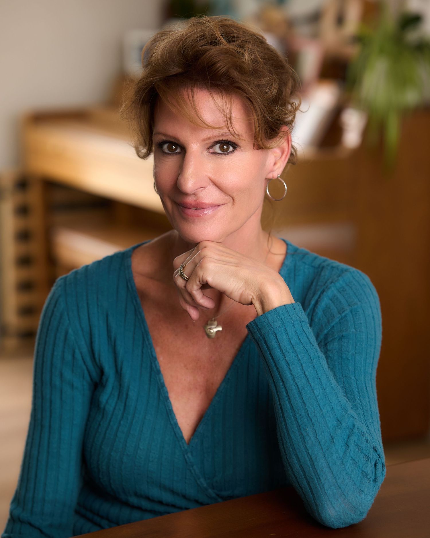 A smiling woman with auburn hair wearing a teal sweater and gold earrings poses with her chin resting on her hand.