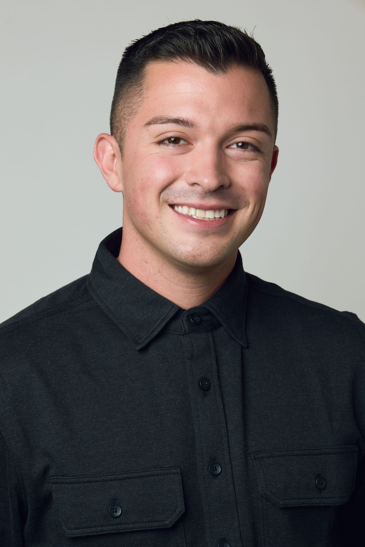 Smiling young man with short dark hair wearing a black button-up shirt against a light gray background.