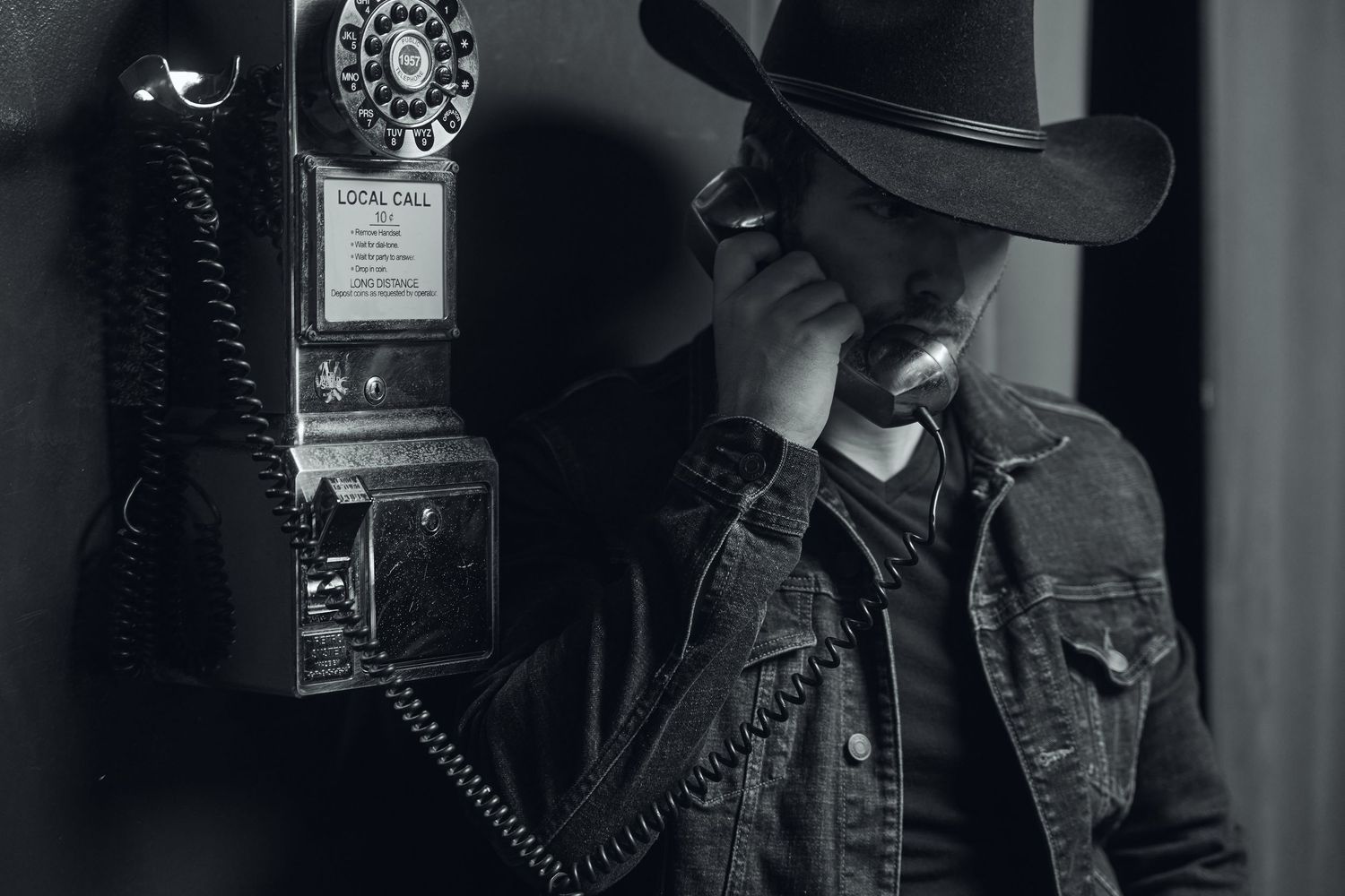 Man in cowboy hat and denim jacket using vintage rotary pay phone, black and white photography.