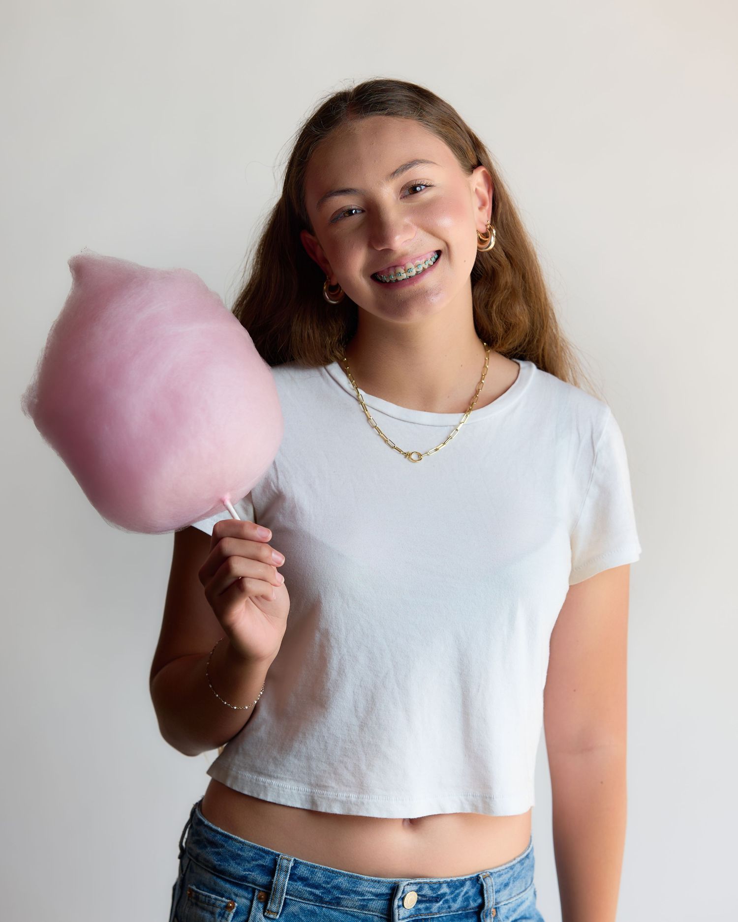 Smiling teenage girl with braces holding pink cotton candy, wearing white crop top and gold necklace.