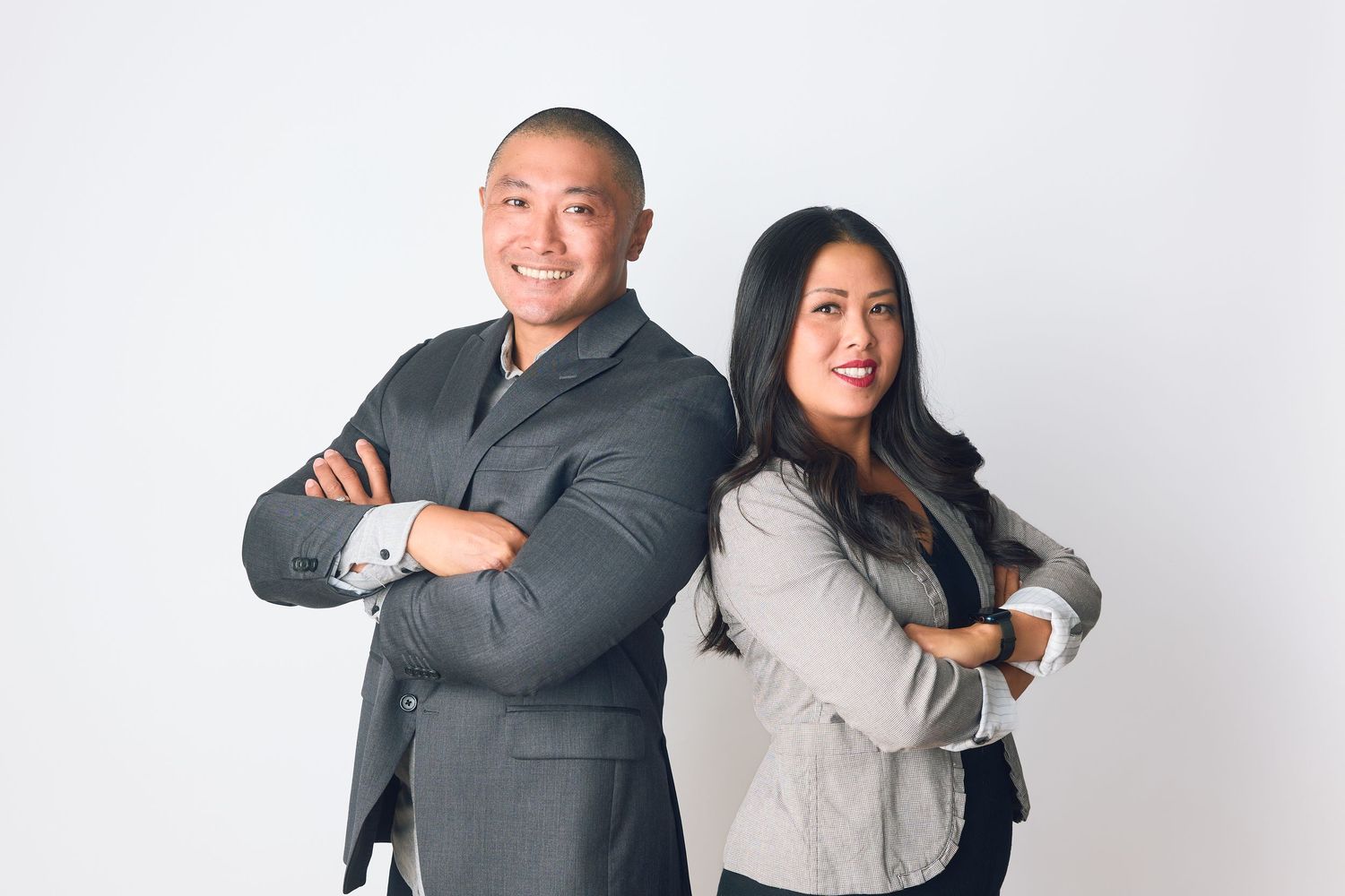 Two business professionals in suits standing back-to-back with arms crossed, smiling confidently against a white background.