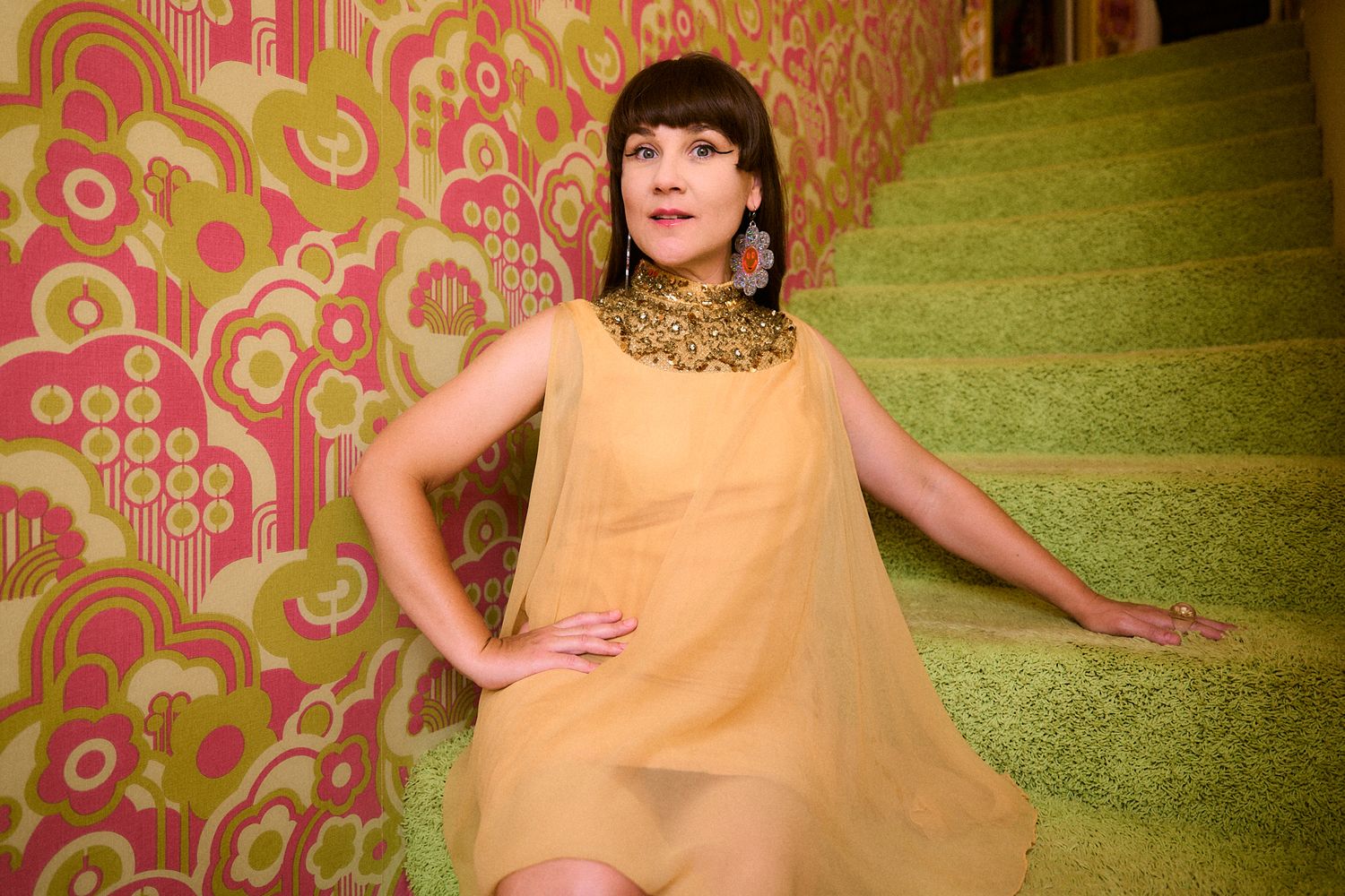 Figure in flowing yellow dress poses against retro pink and orange patterned wallpaper.