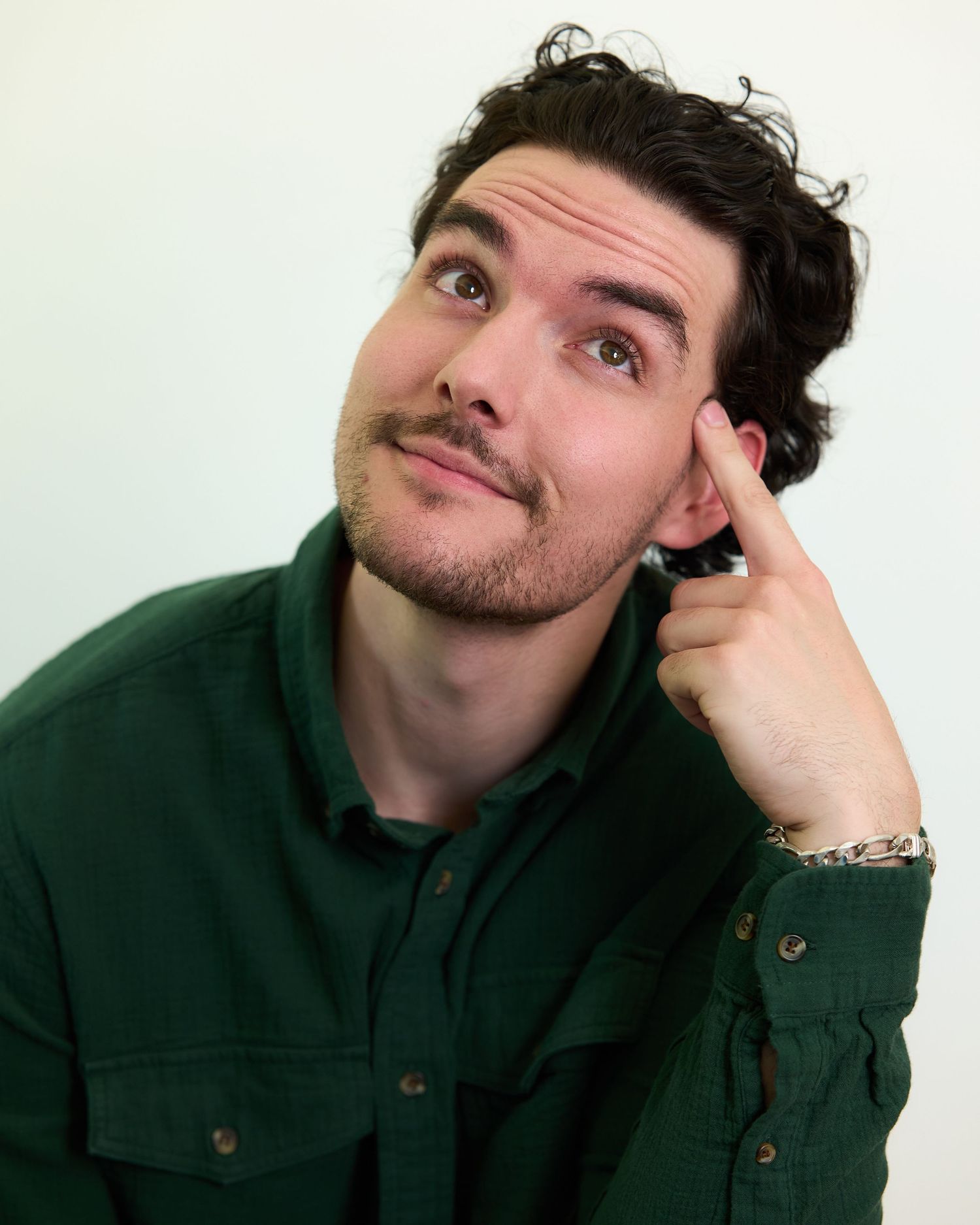 Young man in green shirt posing thoughtfully with finger near temple, looking upward against white background.