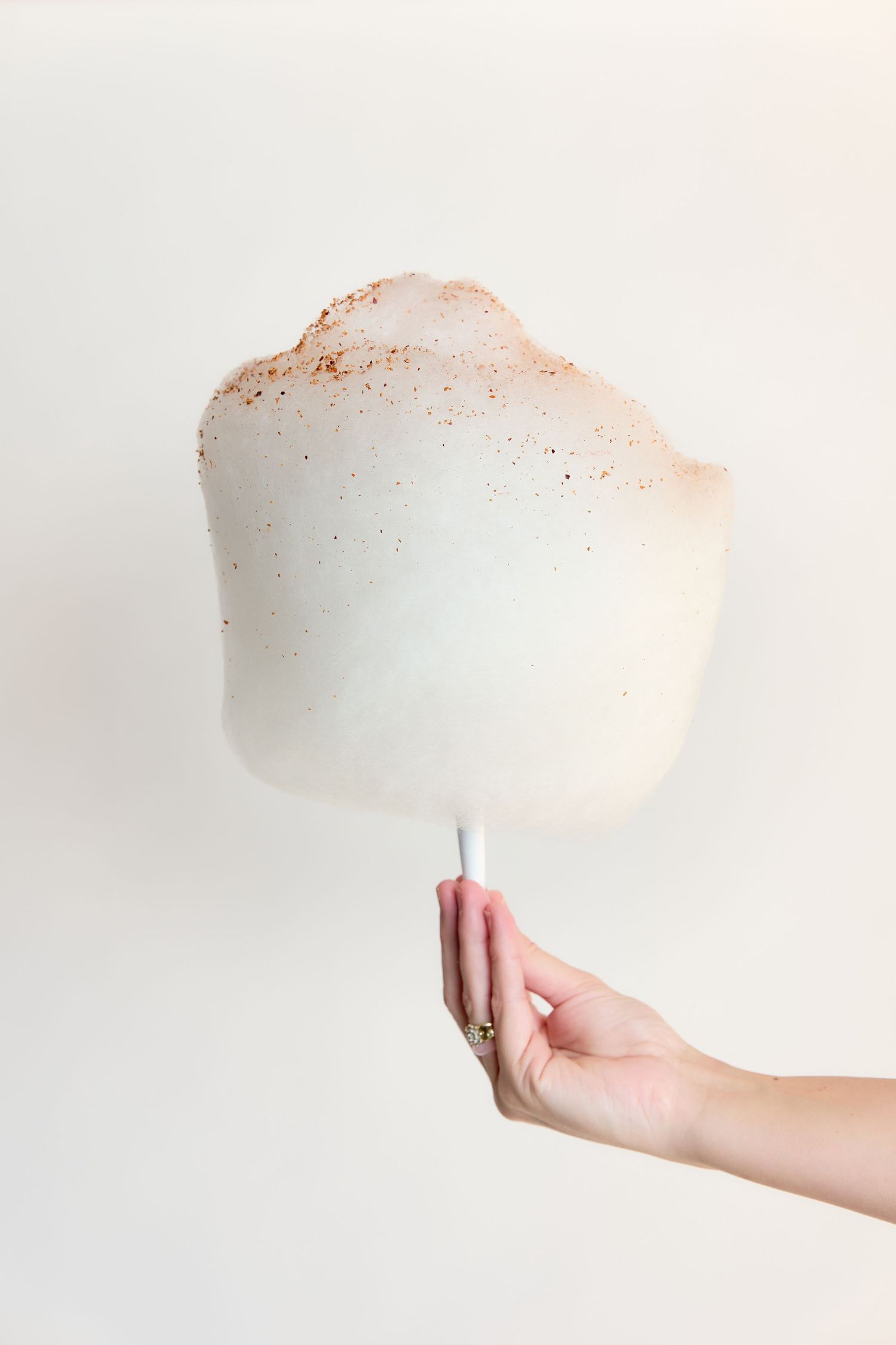 Hand holding a large white cotton candy with cinnamon speckles on a white stick against a cream background.