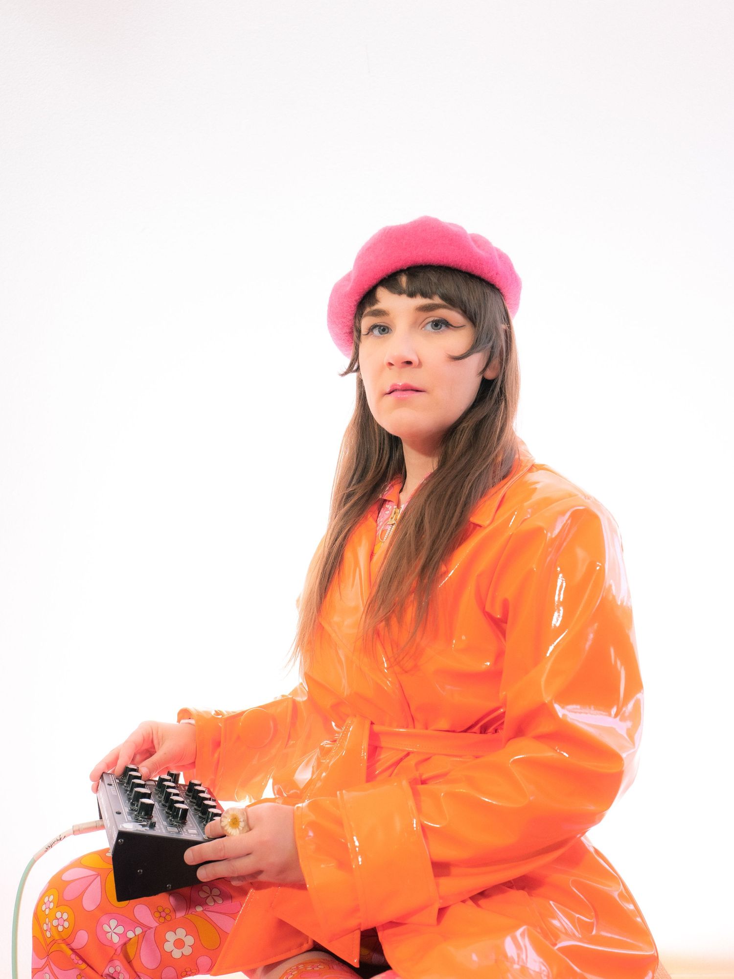 Woman in orange vinyl coat and pink beret holds a synthesizer against a white background.