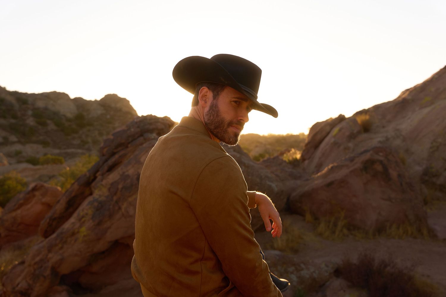 A person in a cowboy hat sits on rocky terrain during golden hour sunset in a desert landscape.