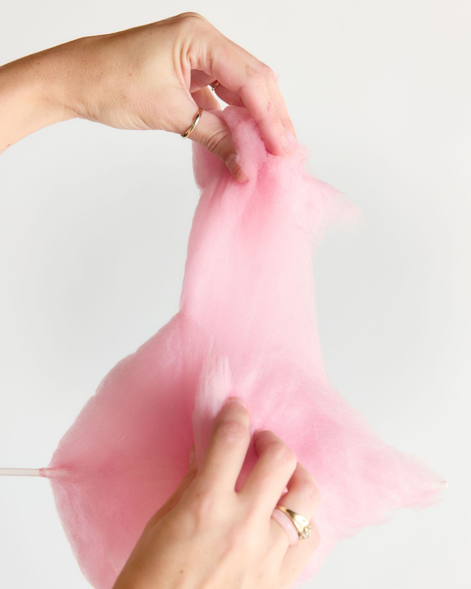 Hands stretching pink cotton candy on a stick against a white background.