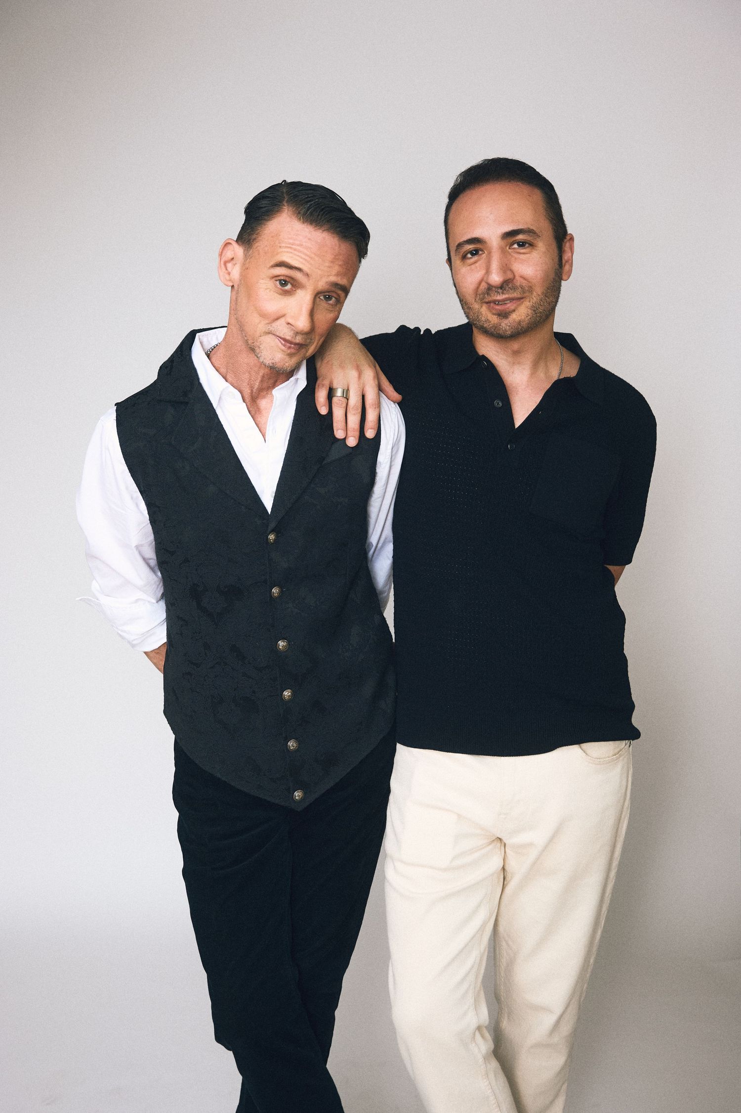 Two stylish men posing together in a studio, one wearing a black vest over white shirt, the other in a black polo.