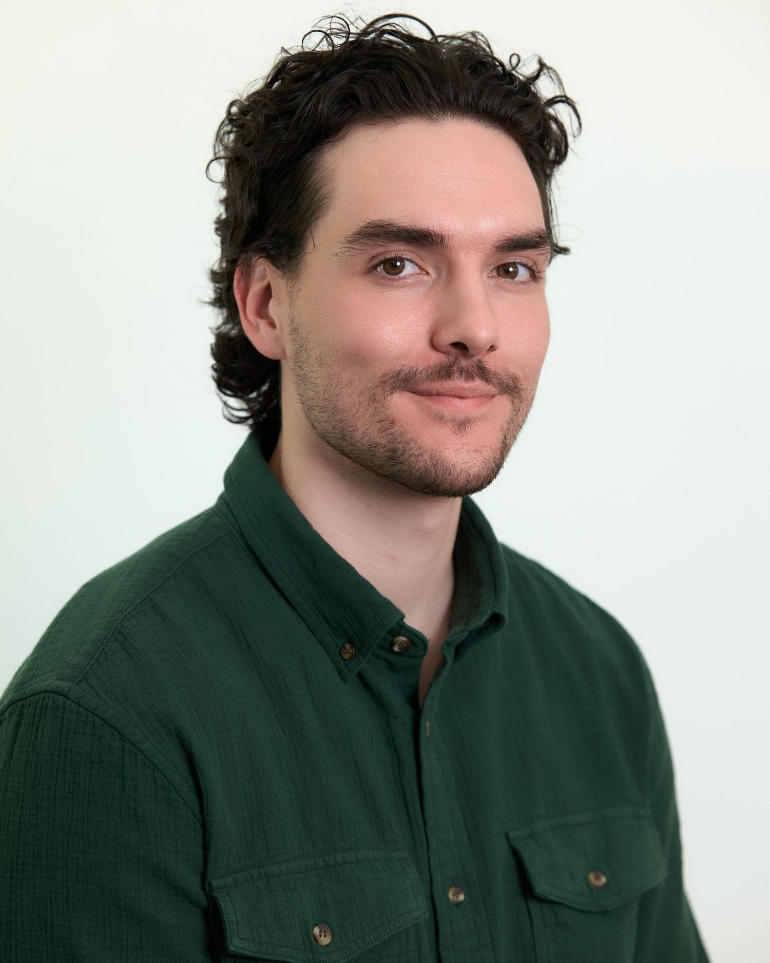 Young man with curly dark hair and beard wearing a dark green button-up shirt poses for a professional headshot.
