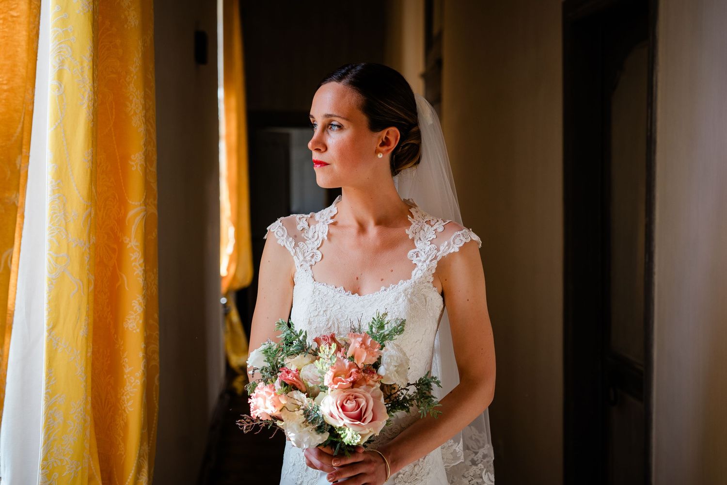 Bride in lace dress holds pastel bouquet while standing by window in warm light.
