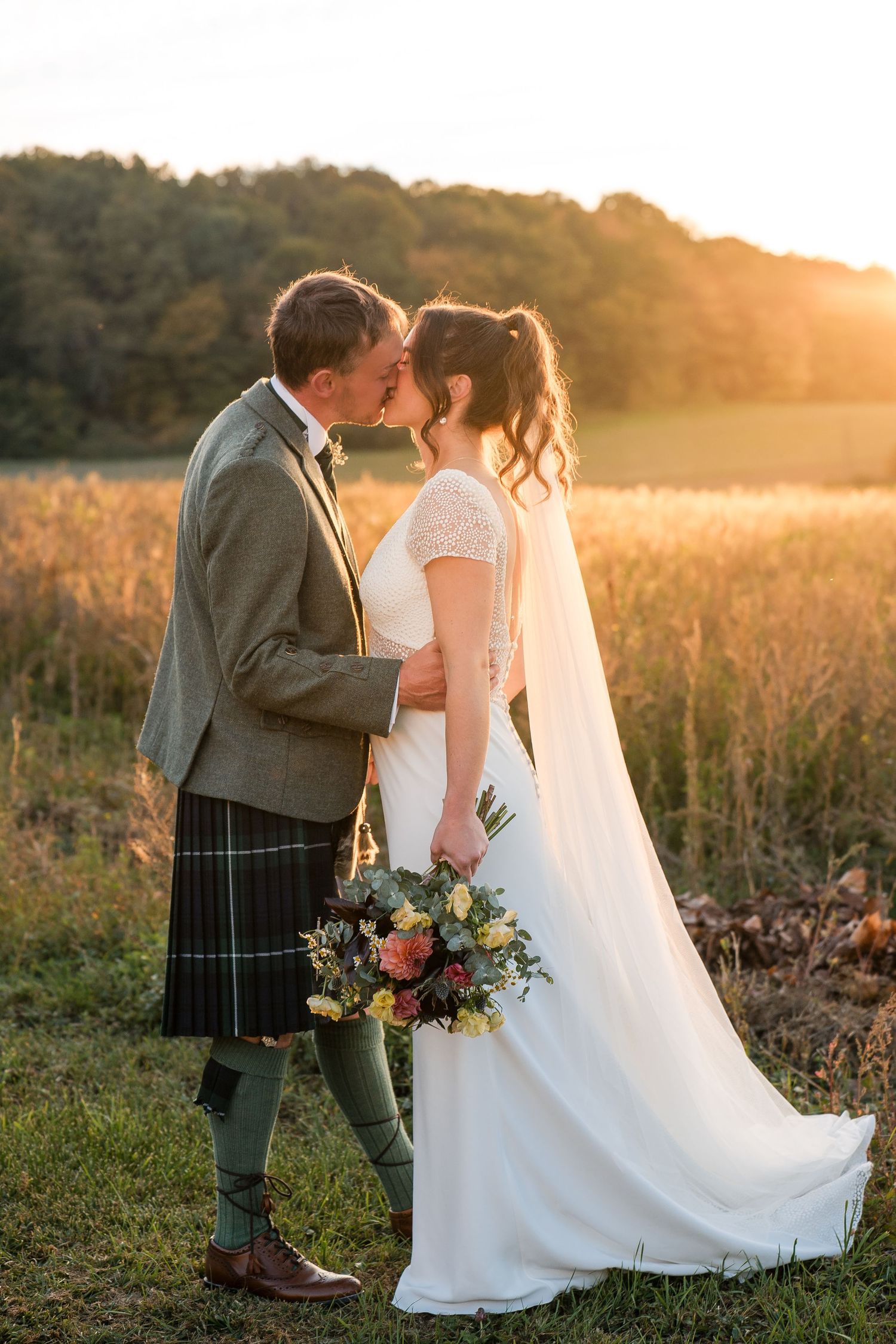 Wedding couple in Scottish attire kiss during golden hour in a meadow with wildflowers.