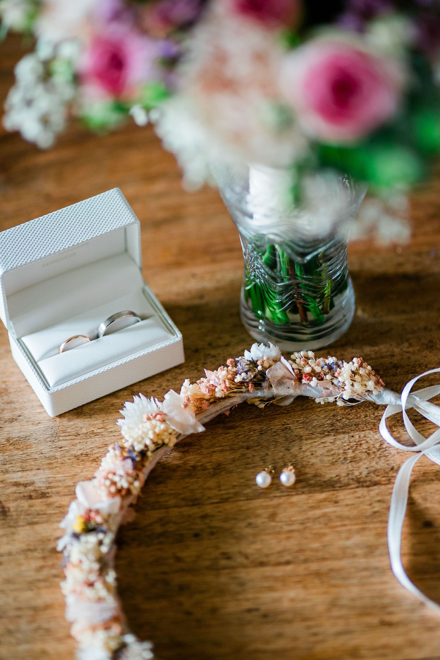 Wedding details flatlay with ring box, floral crown, and pink roses on wooden surface.