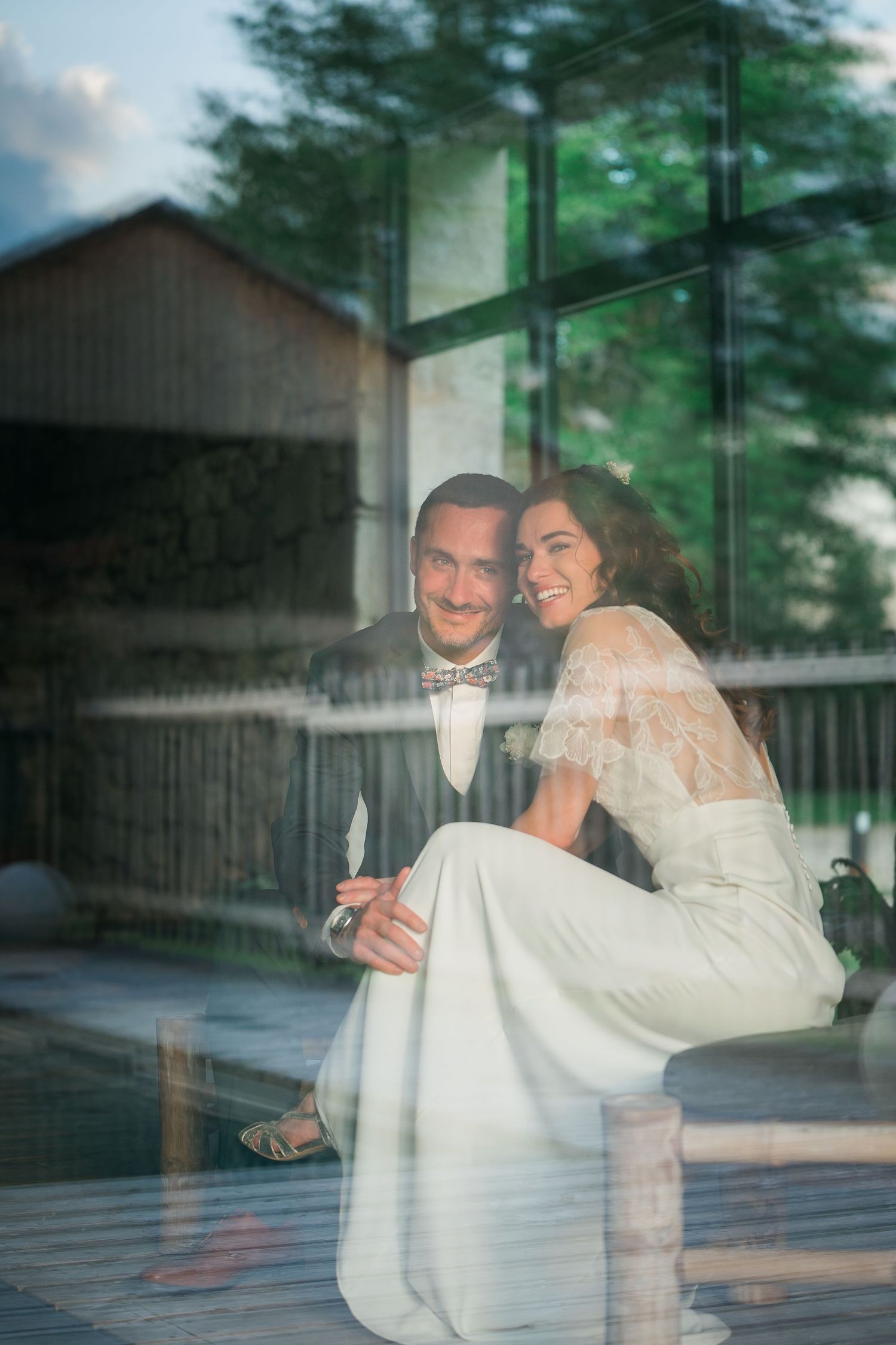 Wedding couple shares intimate moments reflected in glass windows with greenery in background.