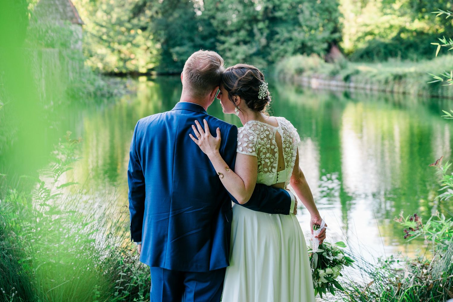 Romantic couple in blue and white attire embrace by a tranquil pond surrounded by lush greenery.