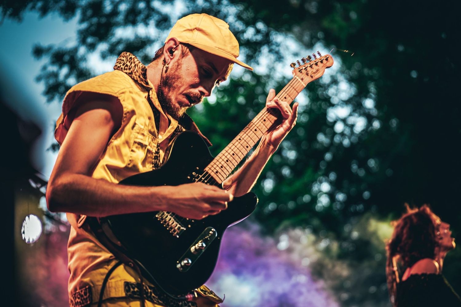 Musician playing electric guitar on stage during live performance with colorful lighting effects in background.